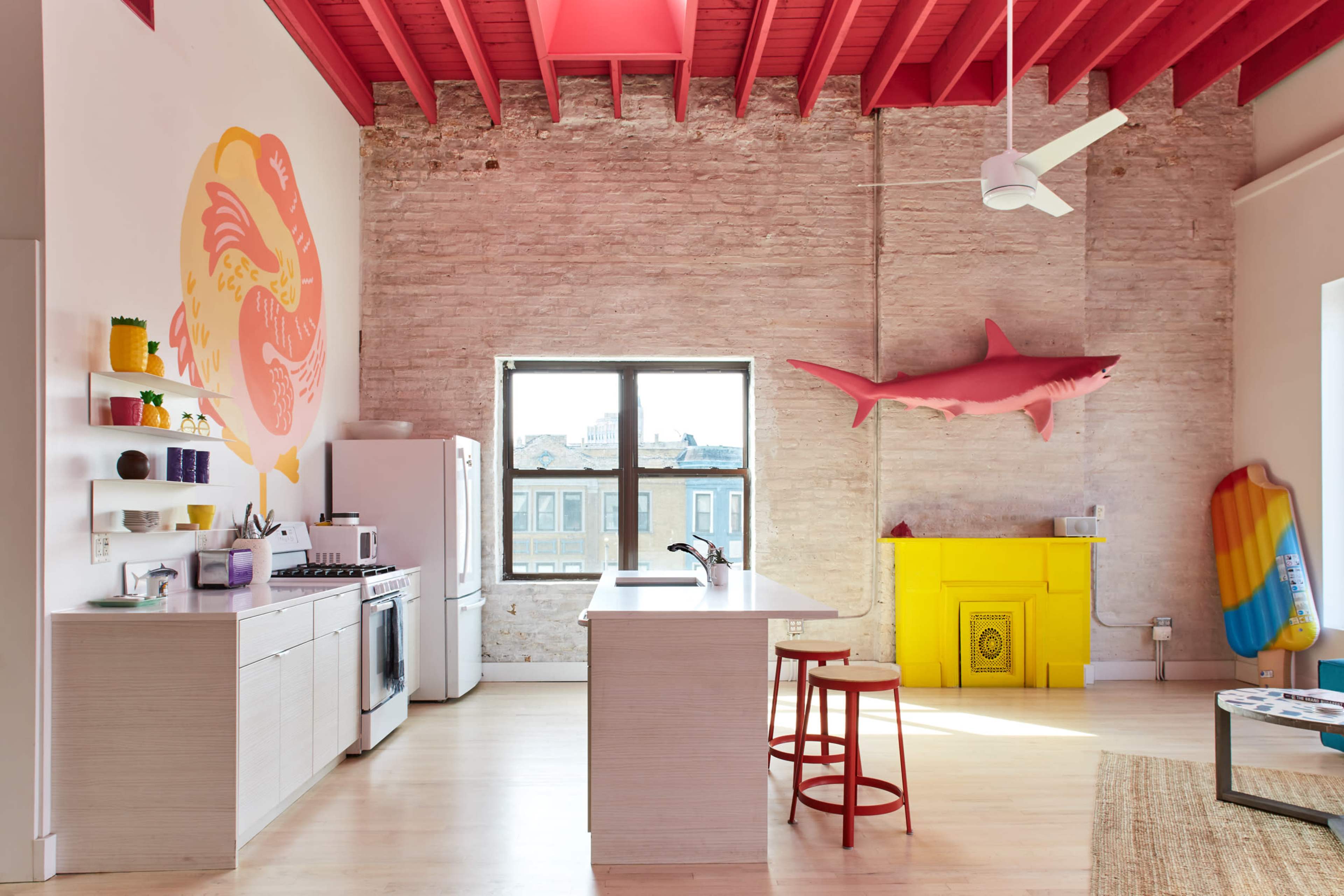 A bright, modern kitchen features a large window, colorful wall art, a yellow accent cabinet, and a surfboard leaning against the wall.