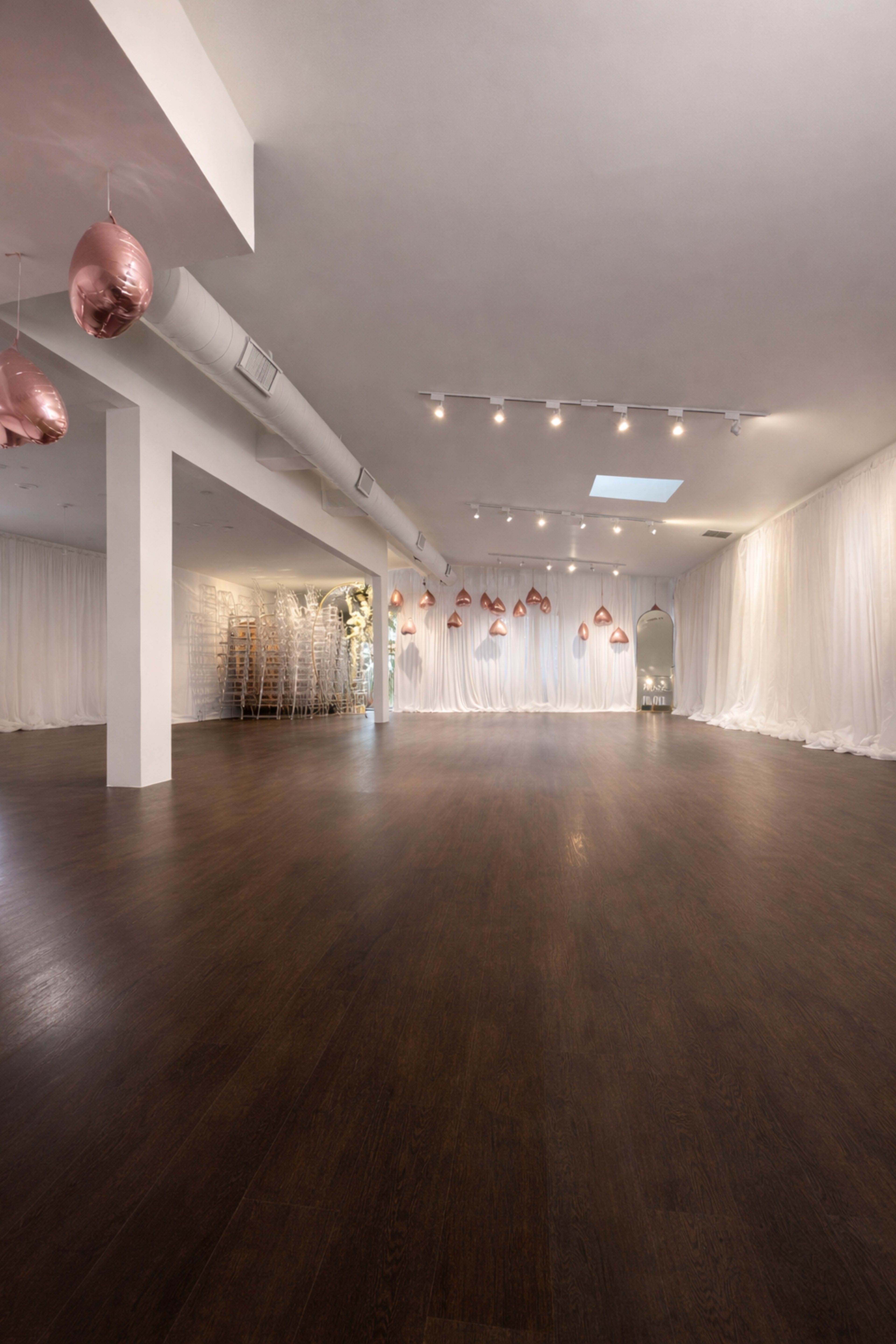 The image shows a spacious, well-lit room with hardwood flooring, white draped fabric along the walls, and decorative rose gold balloons suspended from the ceiling.
