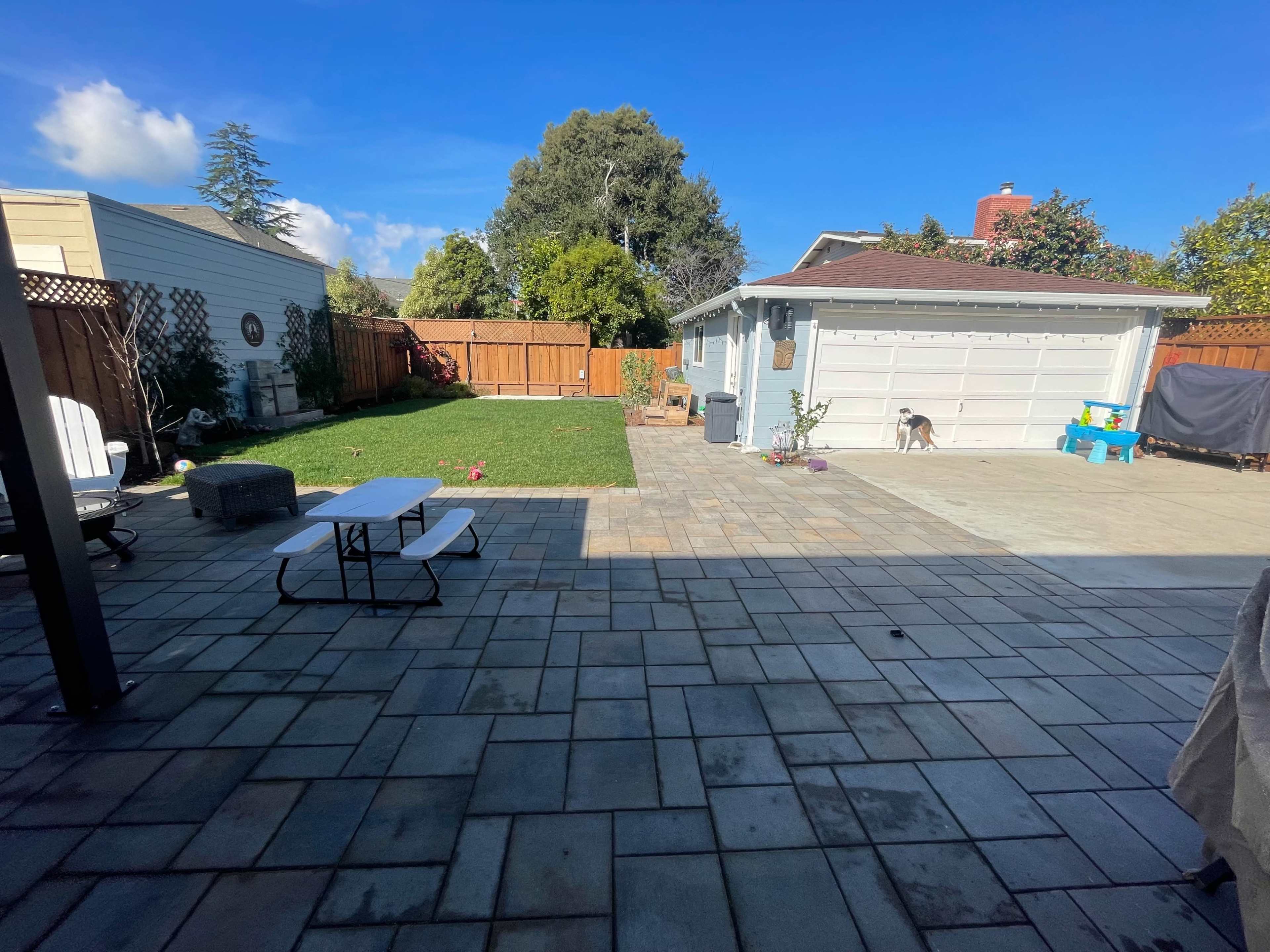 The image shows a backyard with a paved patio, a grassy area, and a garage, surrounded by wooden fencing and a clear blue sky.