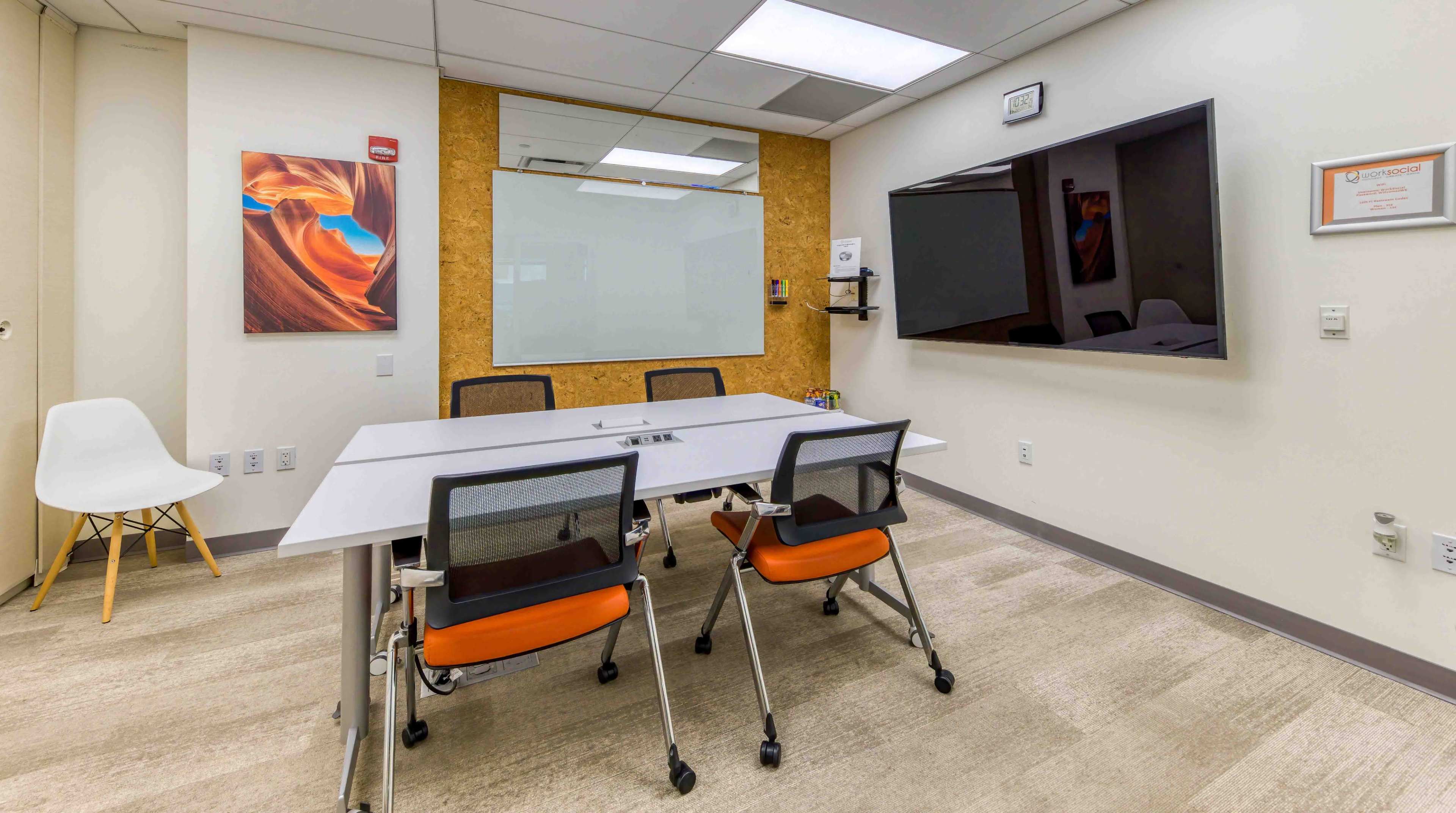 A modern conference room features a white table surrounded by four chairs, a large screen on the wall, a glass whiteboard, and artwork on display.
