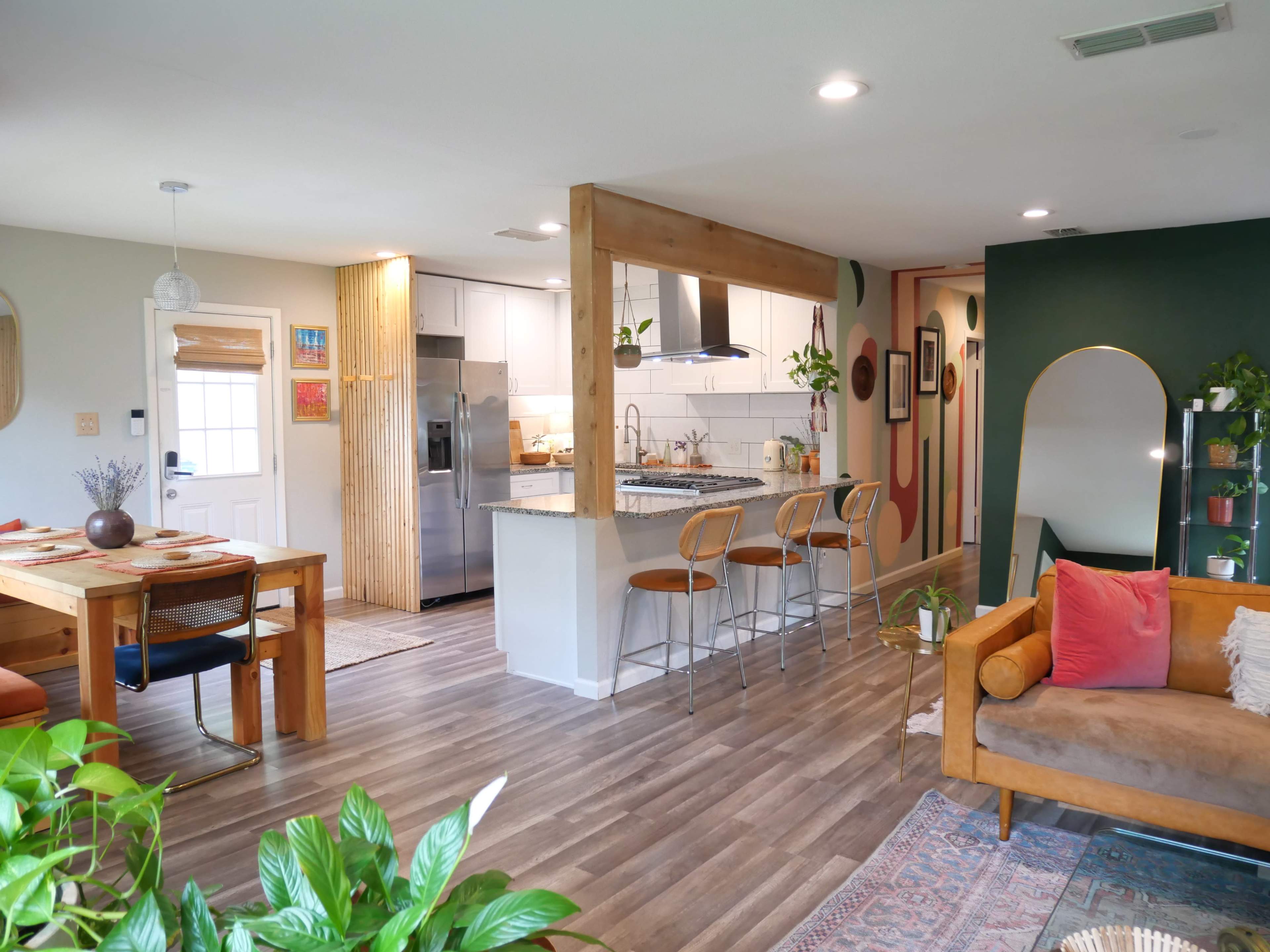 A modern open-concept living space featuring a dining area with a wooden table, a kitchen with bar stools, and a cozy seating area with plants and decorative elements.