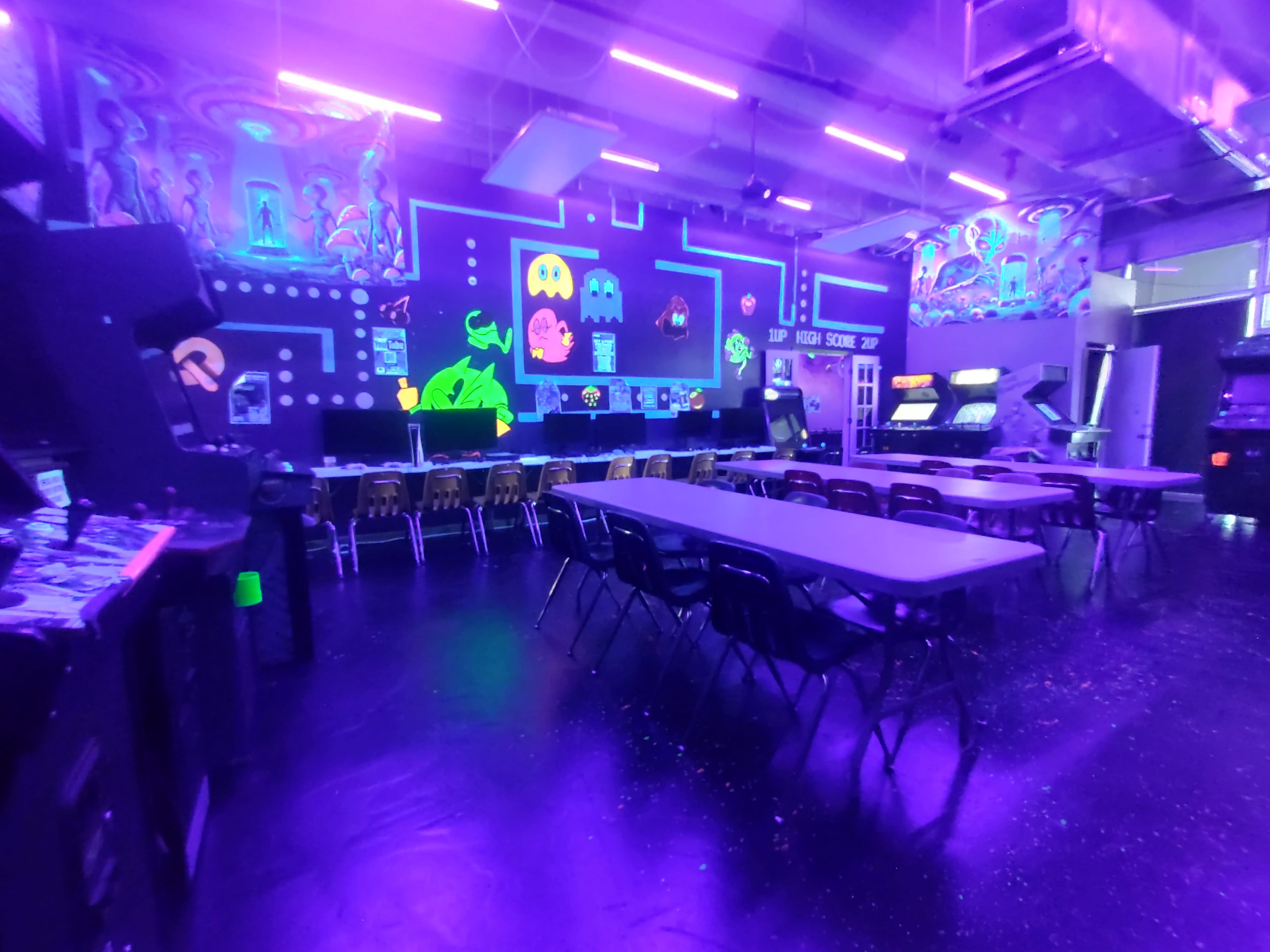 The image features an arcade room with neon lighting, featuring multiple classic arcade machines and tables arranged in a spacious layout.