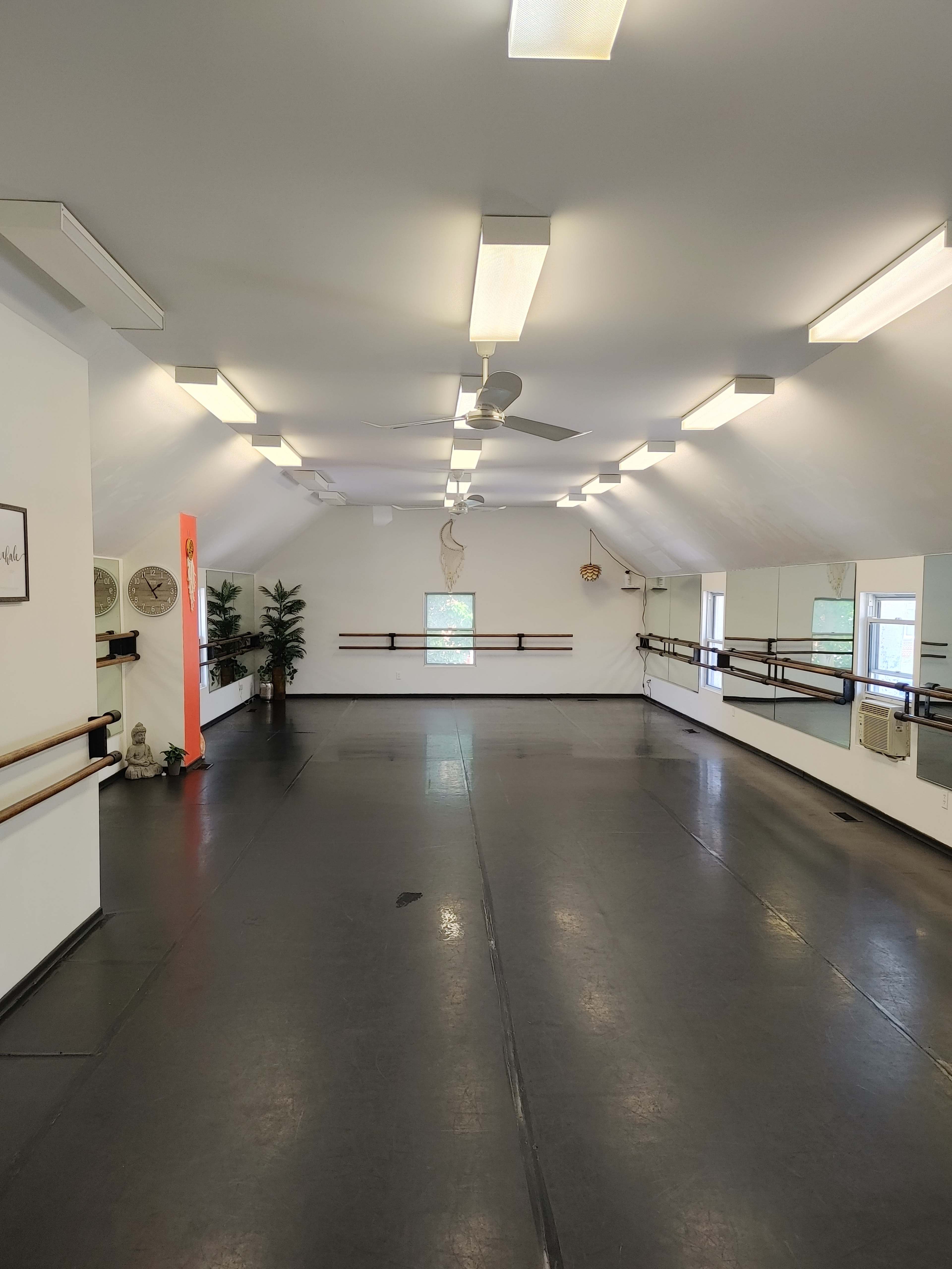 A spacious dance studio with mirrored walls, wooden barre along one side, and bright overhead lights.