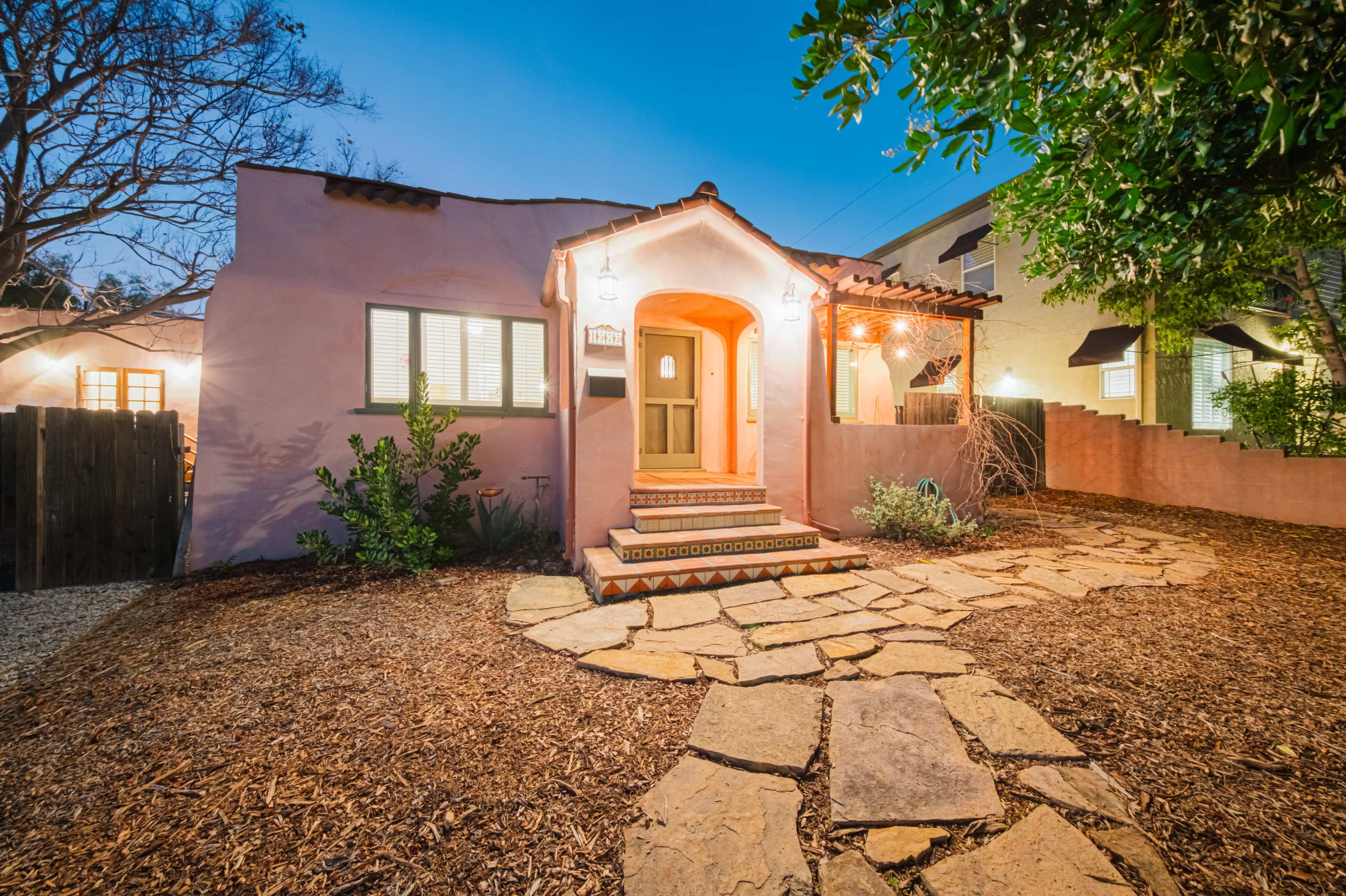 A charming pink stucco house with a tiled entryway is surrounded by a stone pathway and landscaped greenery at dusk.