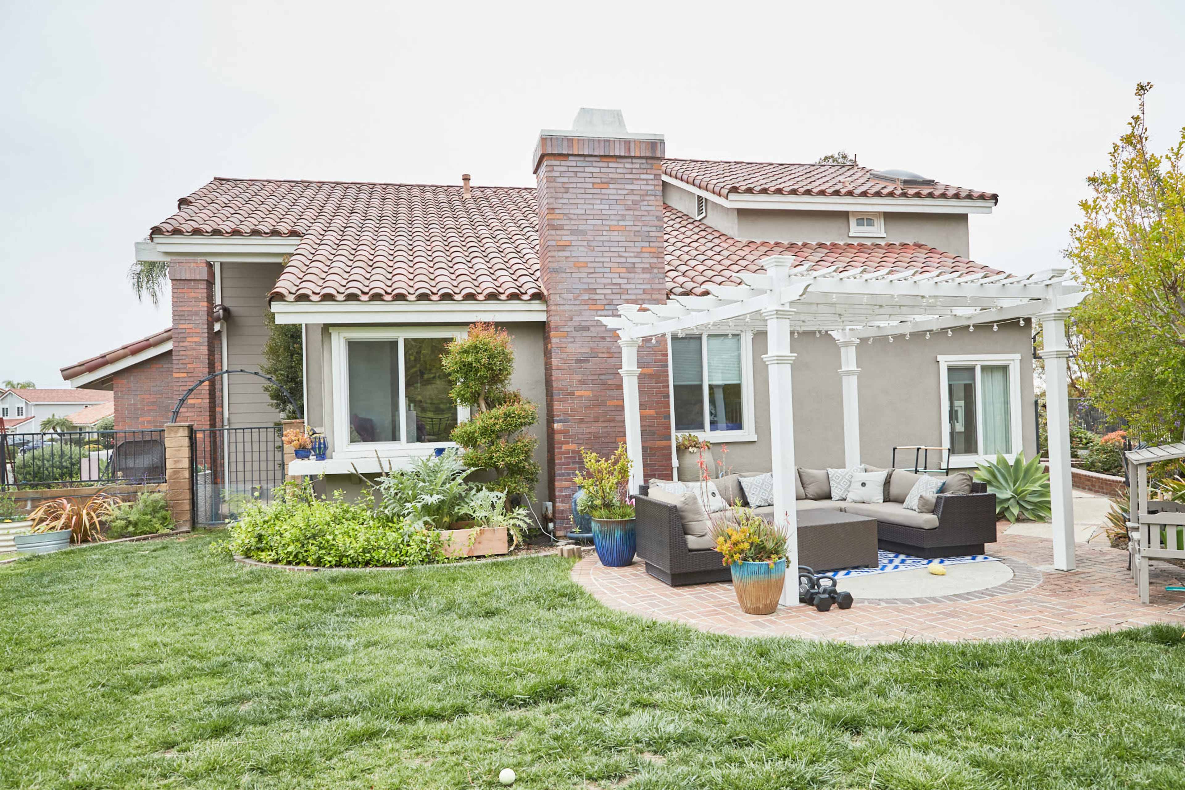 The image shows a modern house with a landscaped backyard featuring a pergola over a seating area and a well-maintained lawn.