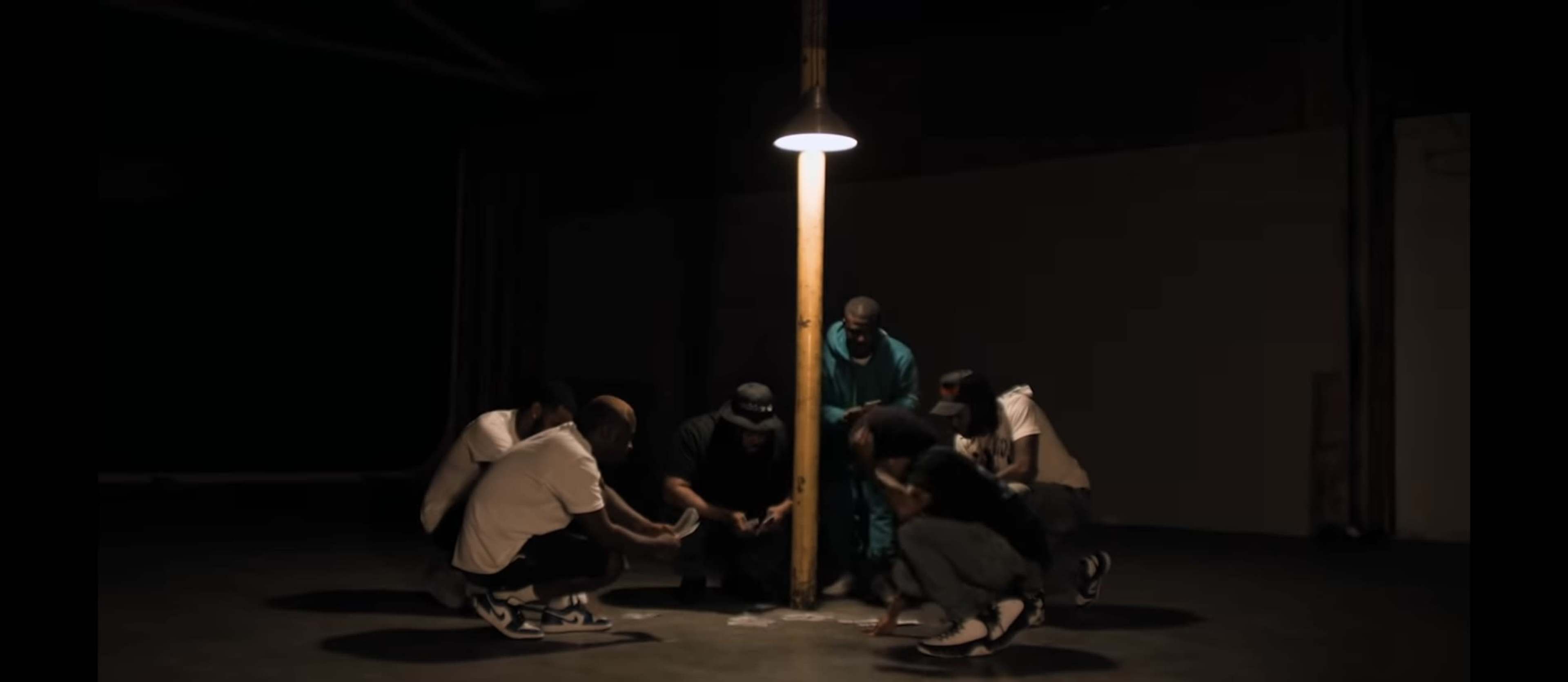A group of six individuals crouches around a lit lamp post in a dimly lit space.