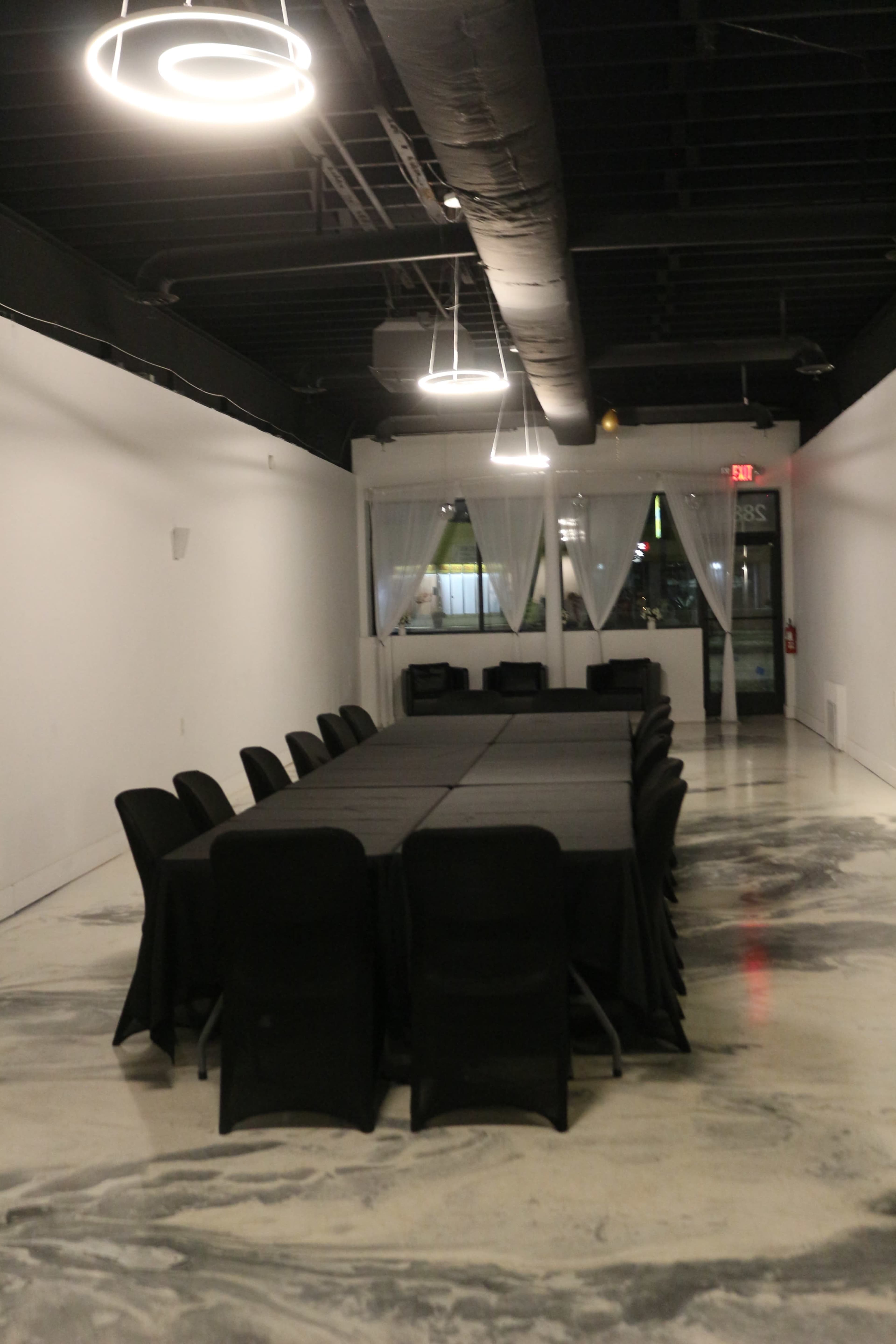 A long, black-covered table is set in a spacious, brightly lit meeting room with black chairs and large windows featuring sheer drapes.