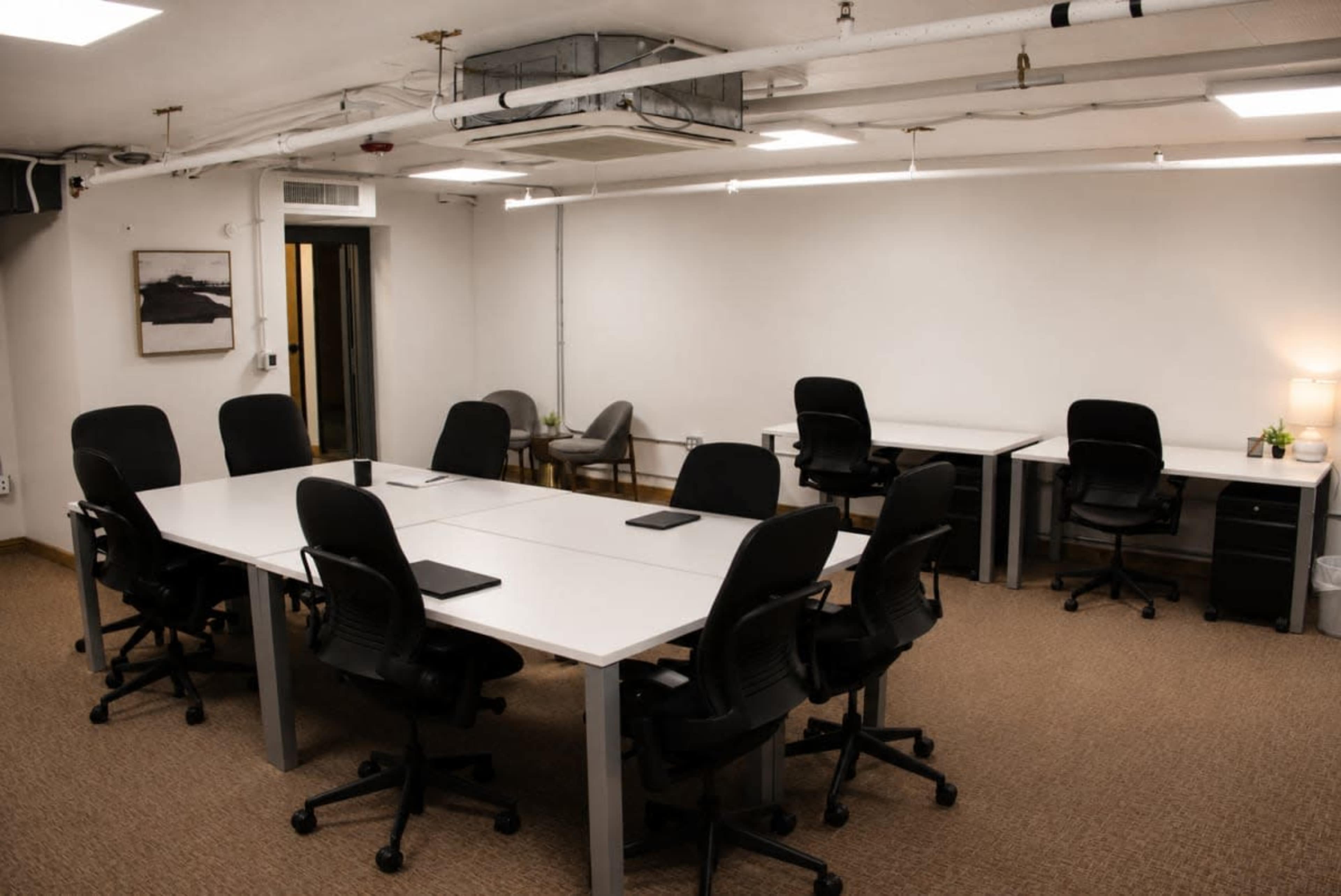 The image shows a modern conference room featuring a long white table surrounded by black office chairs, with additional workstations and minimal decor.