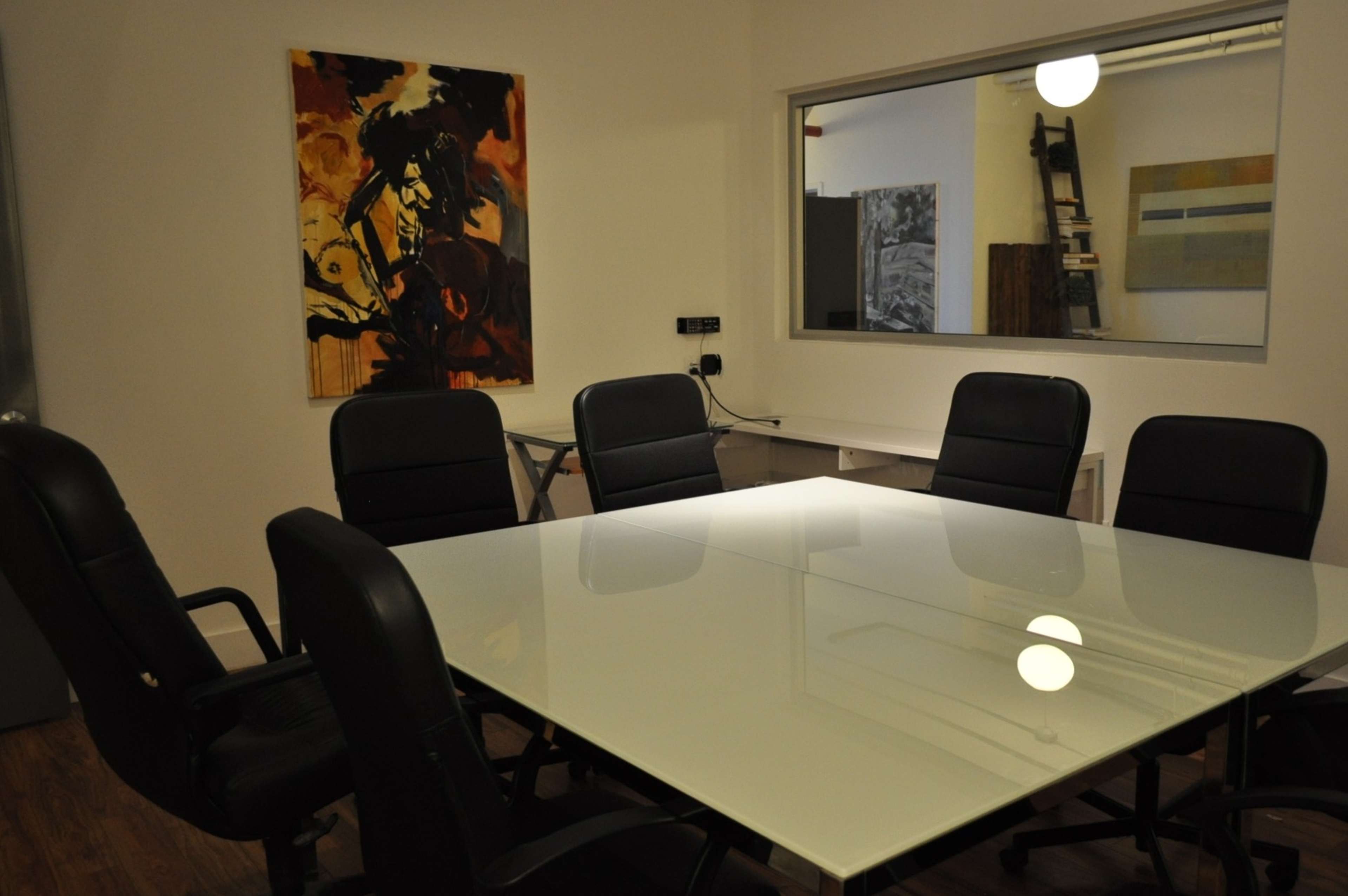 A conference room features a glass table surrounded by black chairs, with artwork on the walls and a large window allowing light to enter.