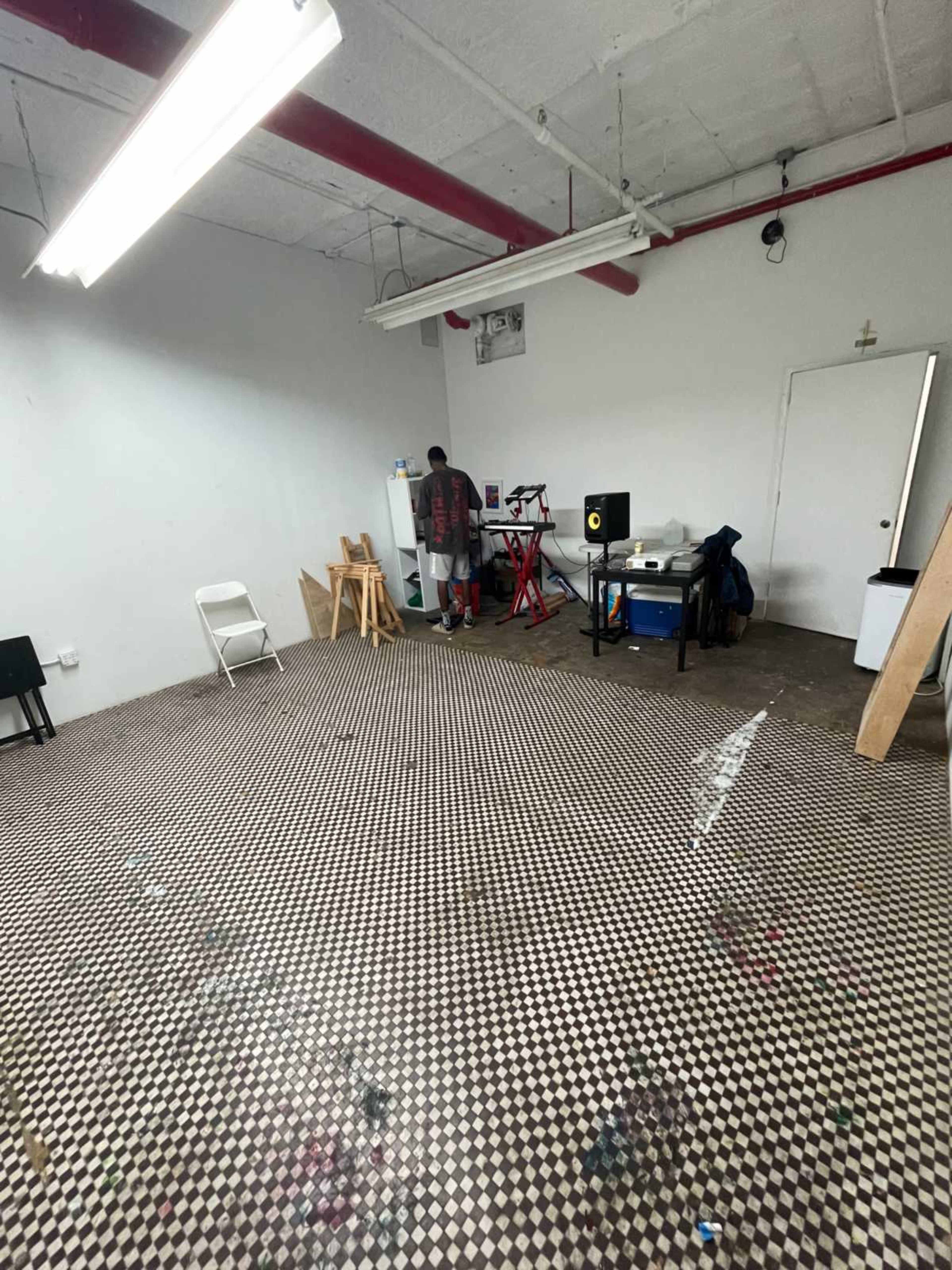 A person stands near a worktable in a spacious, empty room with a checkered floor and minimal furnishings.