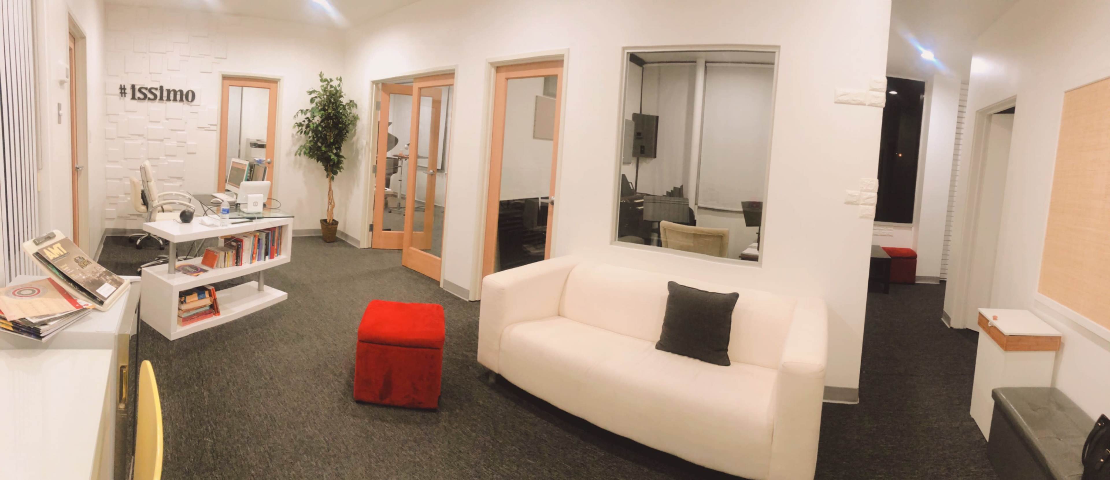 A modern office space with white walls, a white sofa, a red ottoman, and glass-enclosed meeting rooms.