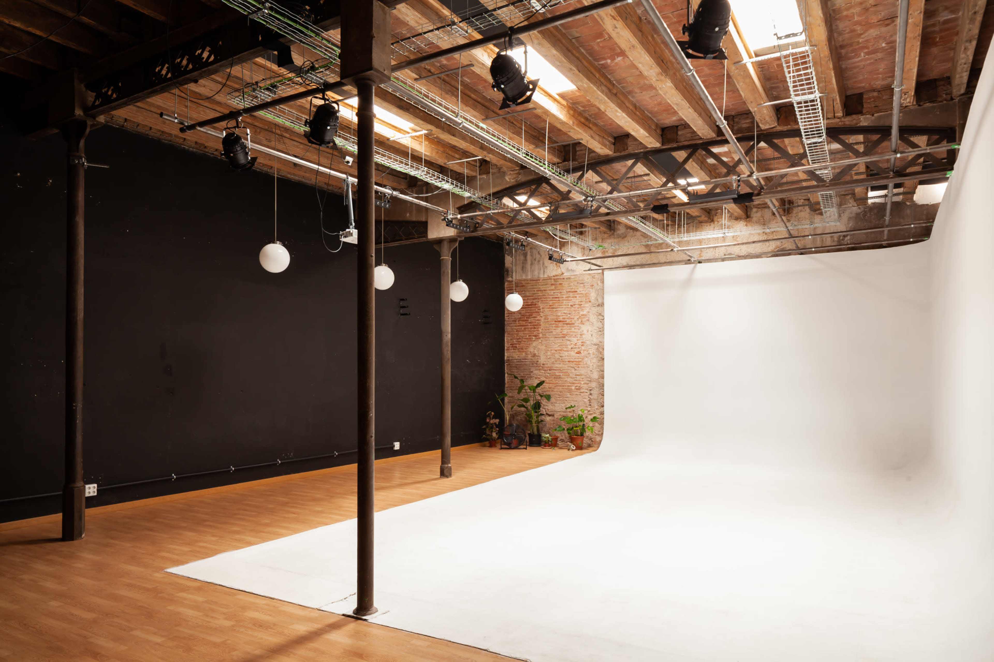 A spacious photography studio with a white backdrop, wooden flooring, exposed brick walls, and overhead lighting.