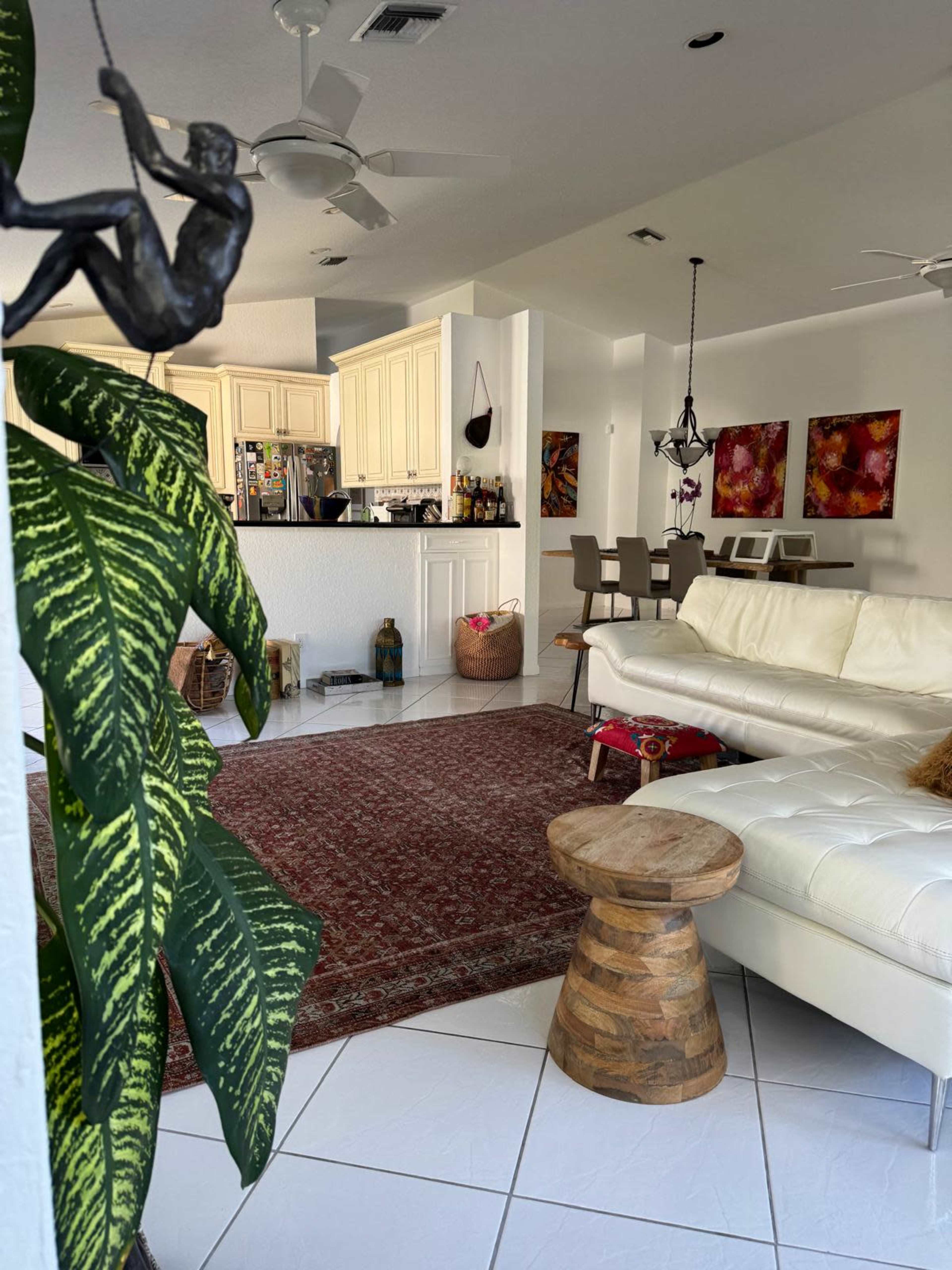 The image shows a spacious living area with a white leather sofa, a wooden stool, a large rug, and modern decor, including a kitchen visible in the background.