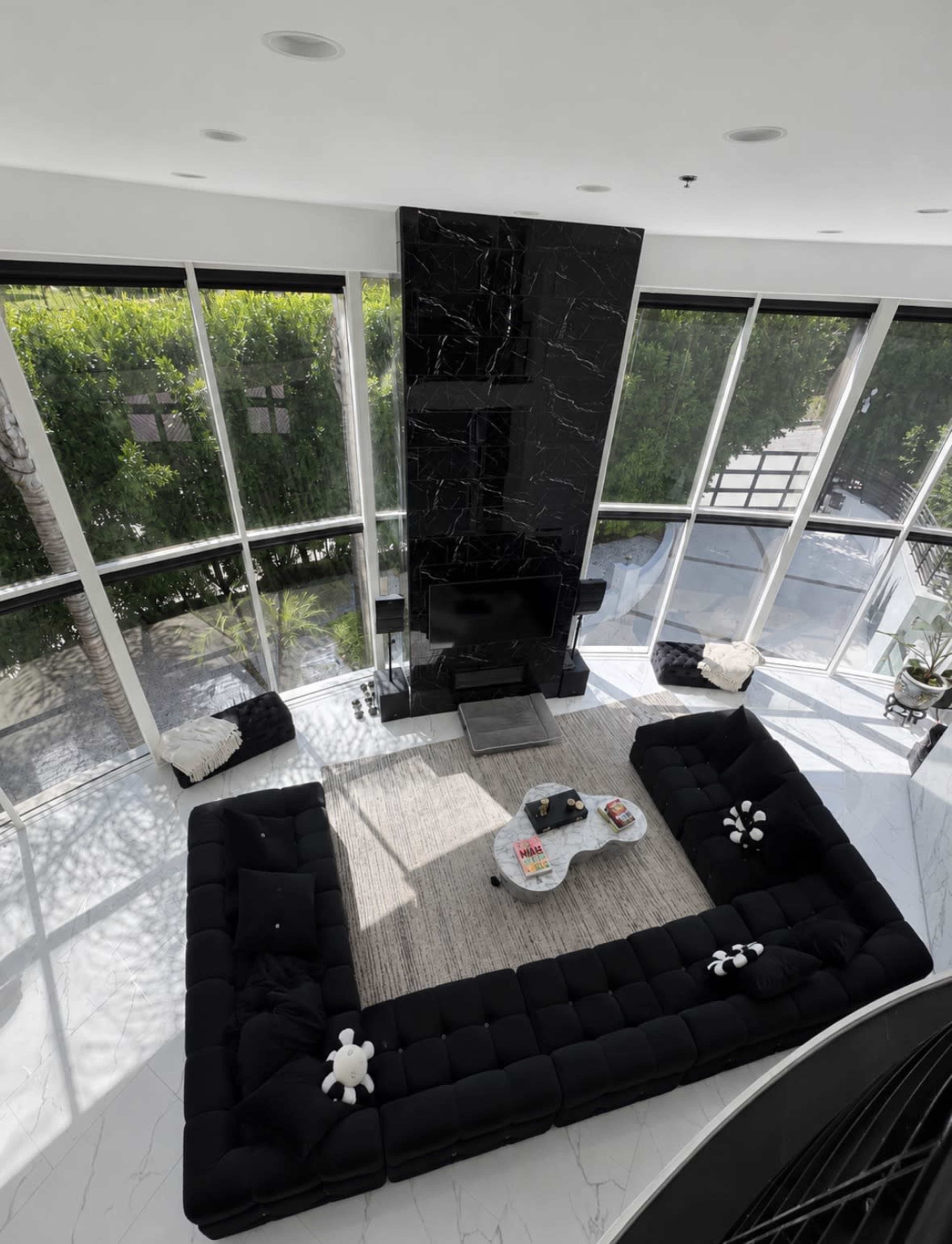 A spacious modern living room features a large black sectional sofa, a central coffee table, and a sleek black marble accent wall, while tall windows offer a view of greenery outside.