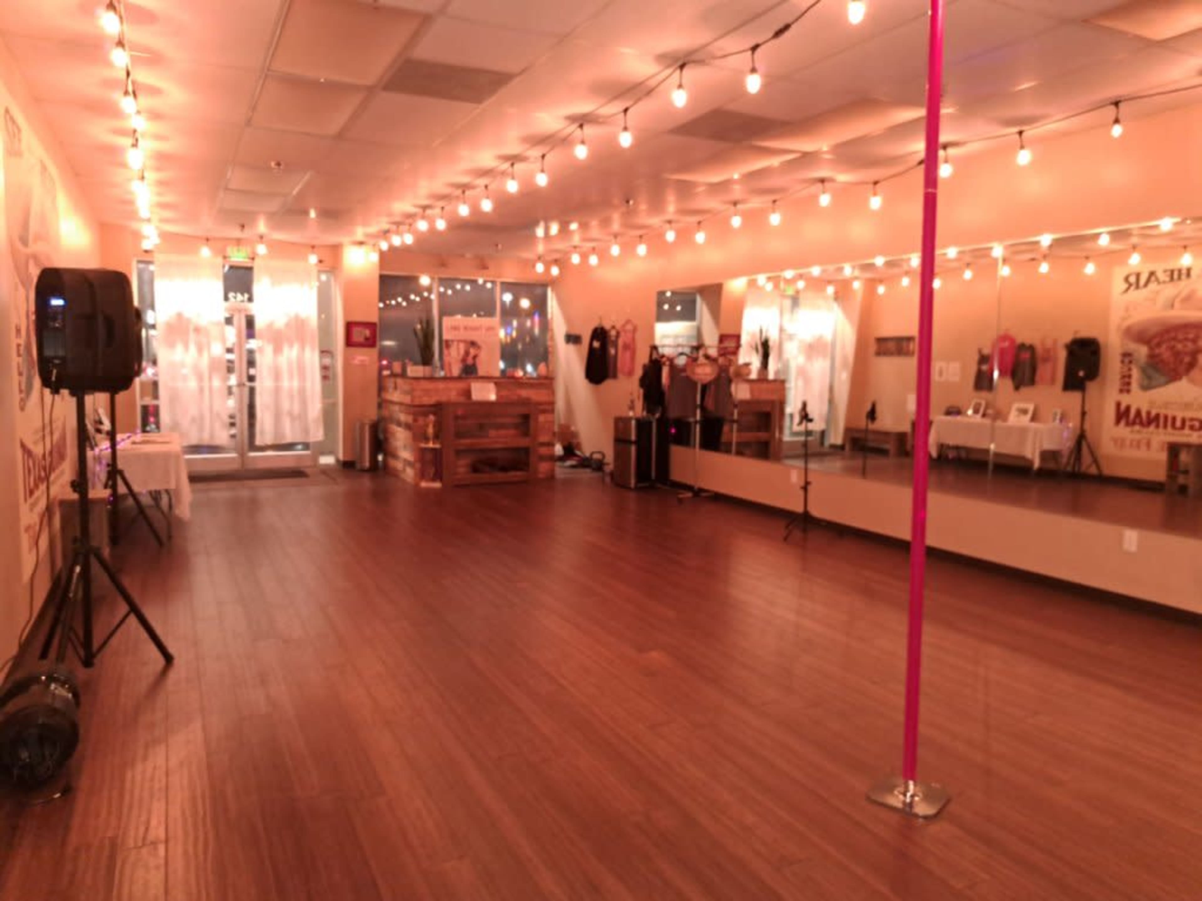 The interior of a dance studio features wooden flooring, a pink pole, mirrored walls, and warm string lights overhead.