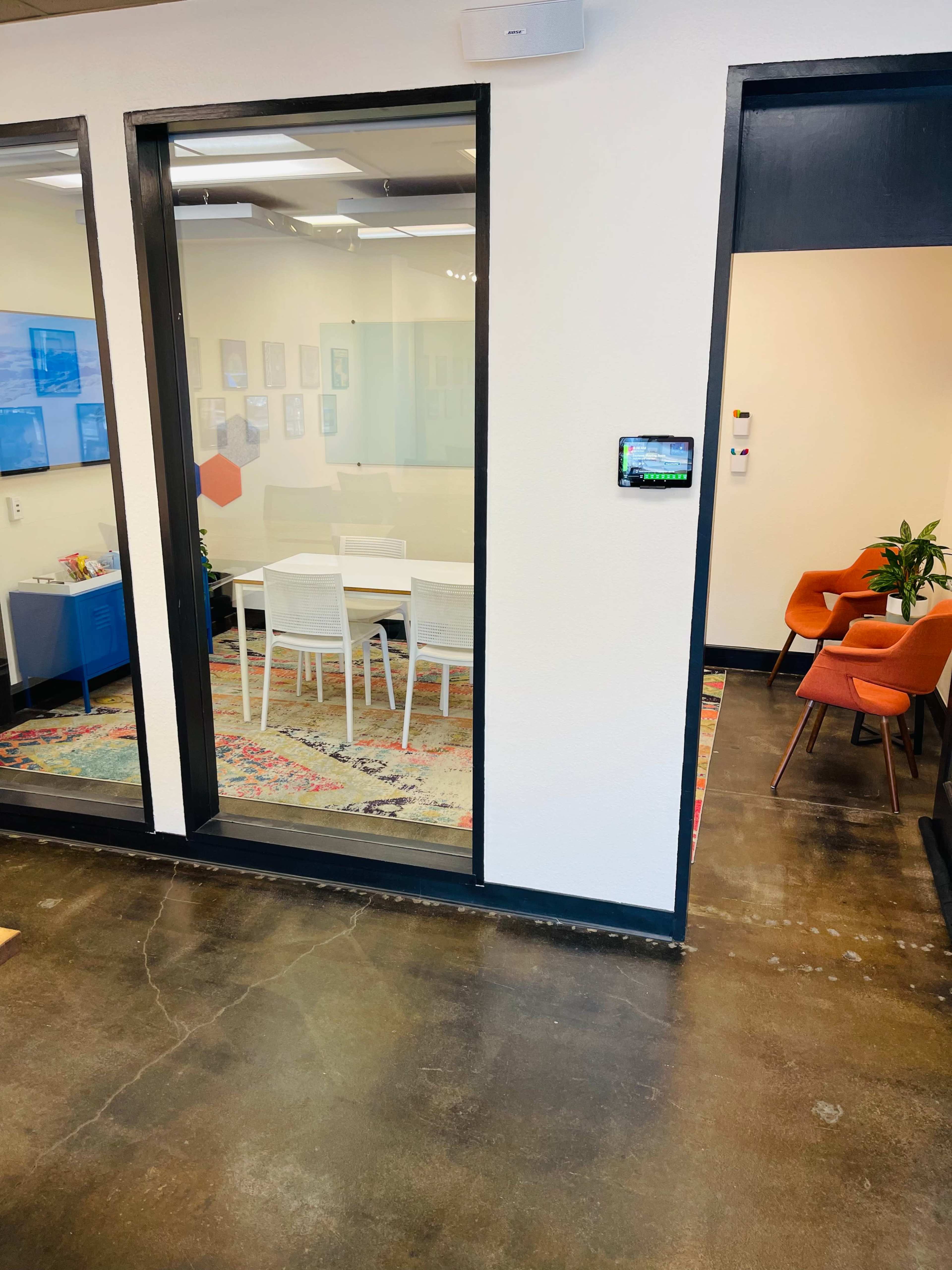 A modern office space featuring a glass-walled conference room with a table and chairs, alongside a cozy seating area with orange chairs and potted plants.