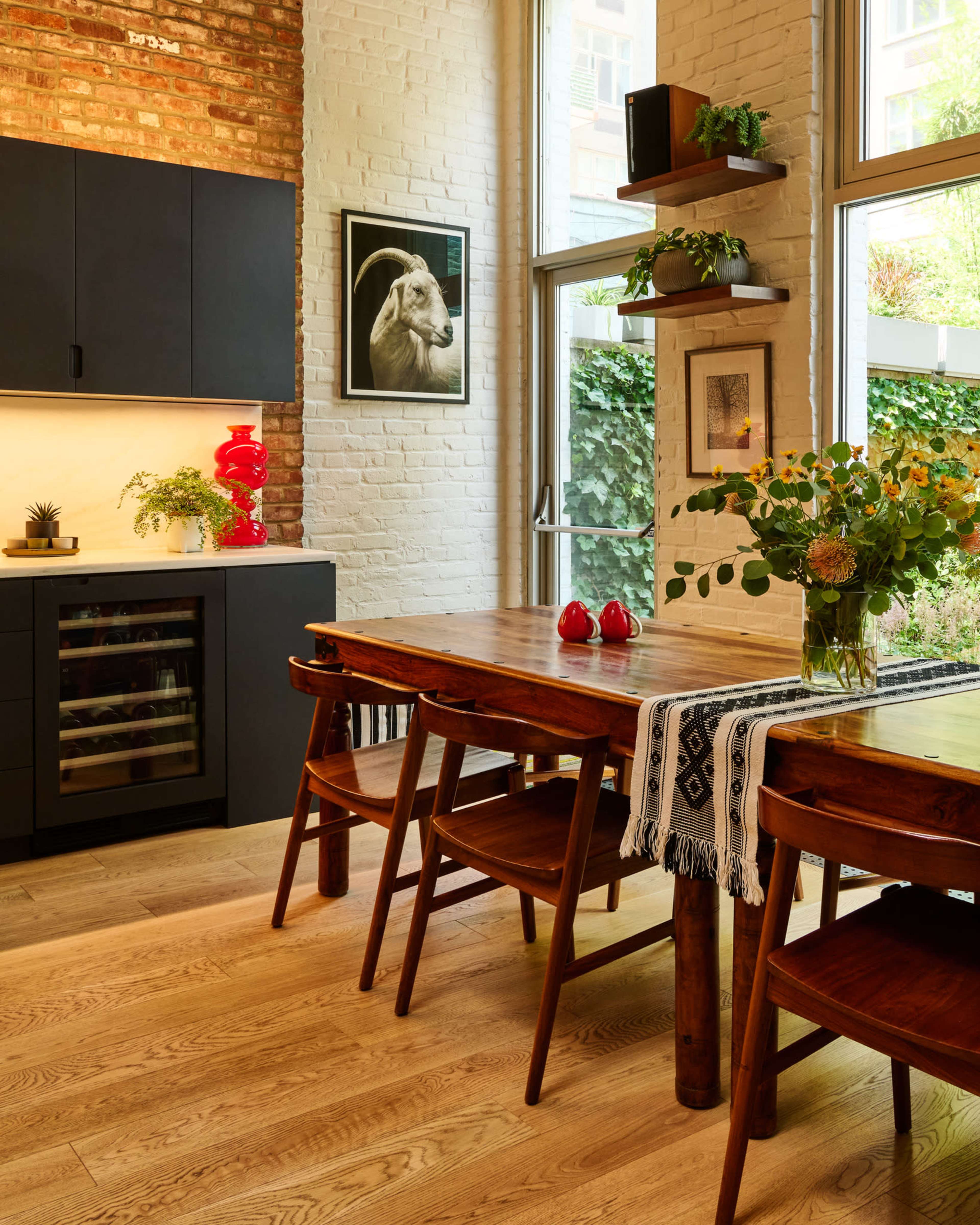 A spacious kitchen and dining area features a wooden table, chairs, and a wall of windows with greenery outside, complemented by black cabinetry and artwork on the walls.