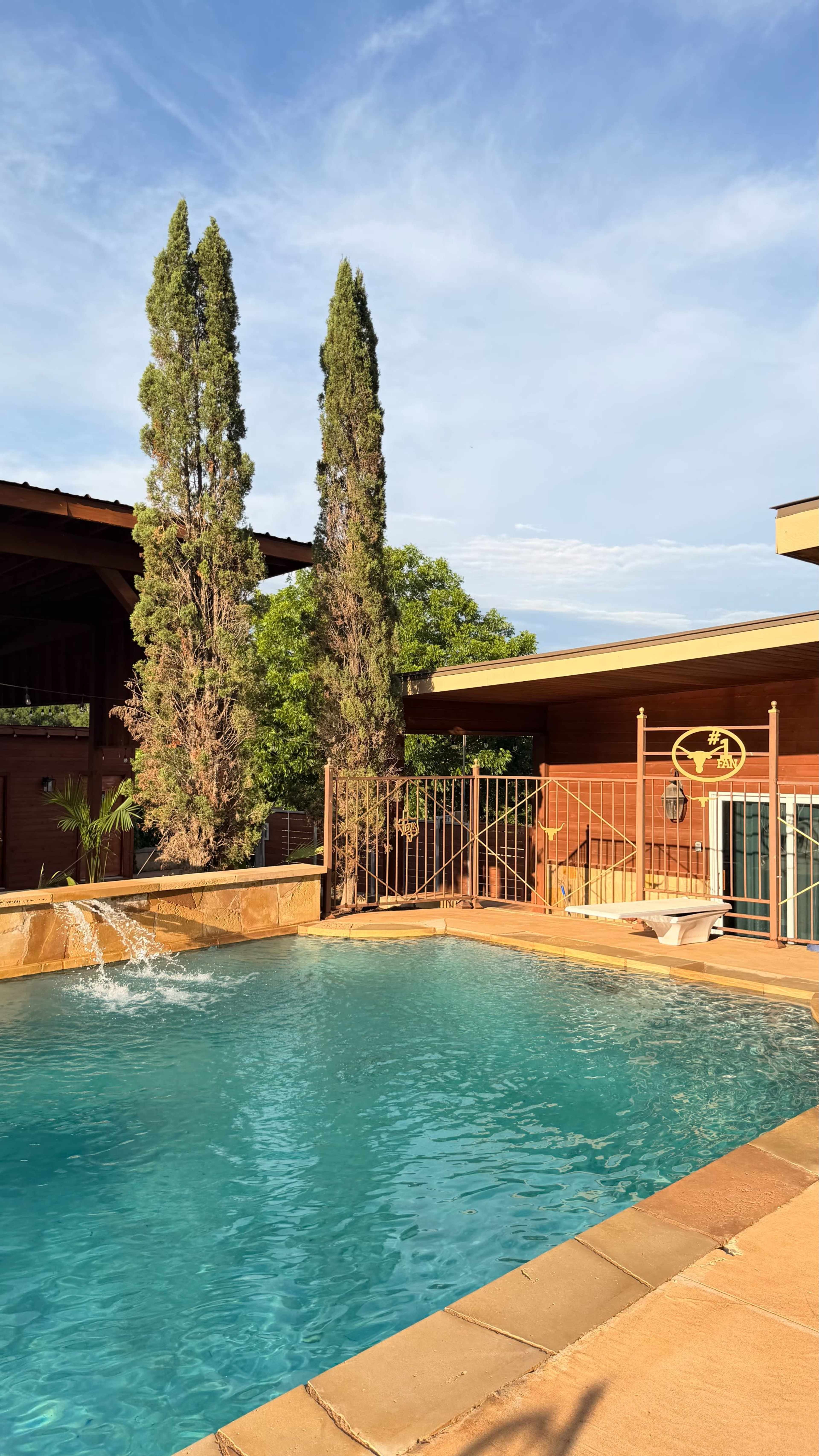 A turquoise swimming pool with a waterfall feature is situated in a backyard surrounded by tall cypress trees and a wooden fence.