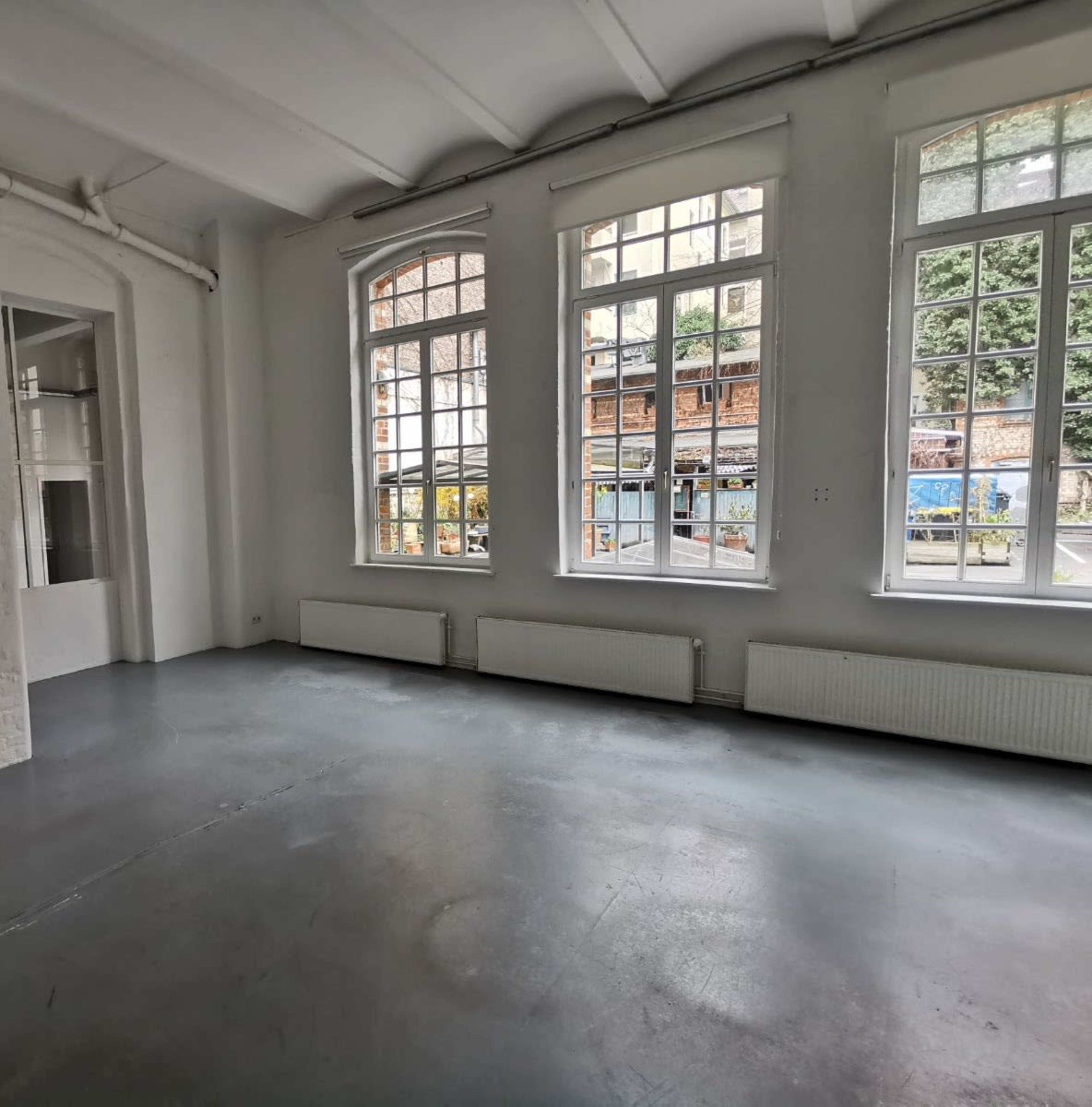 The image shows an empty room with large windows allowing natural light and a view of an outdoor area with buildings and greenery.