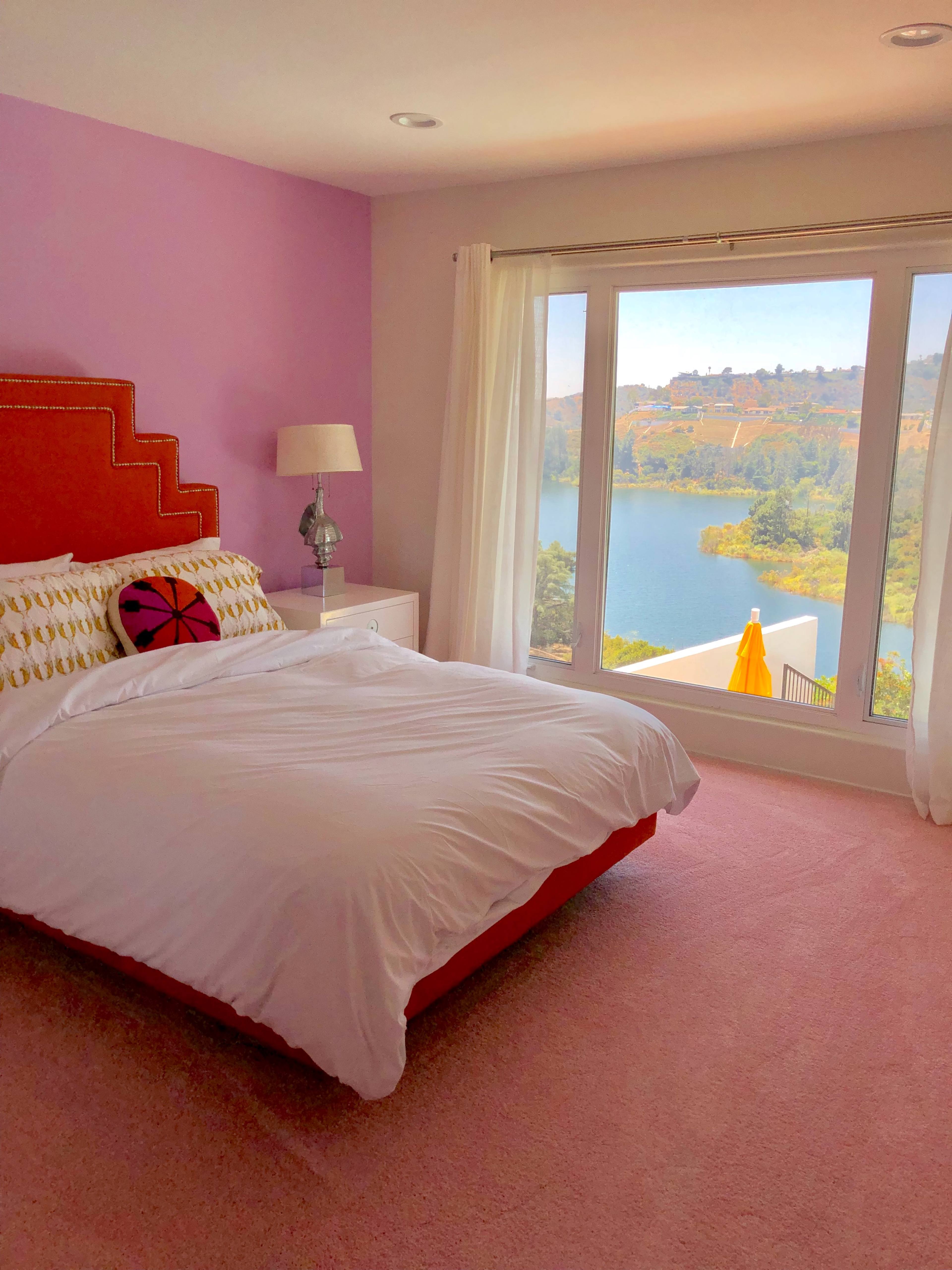 The bedroom features a pink wall and bedding, with a large window overlooking a body of water and a distant landscape.