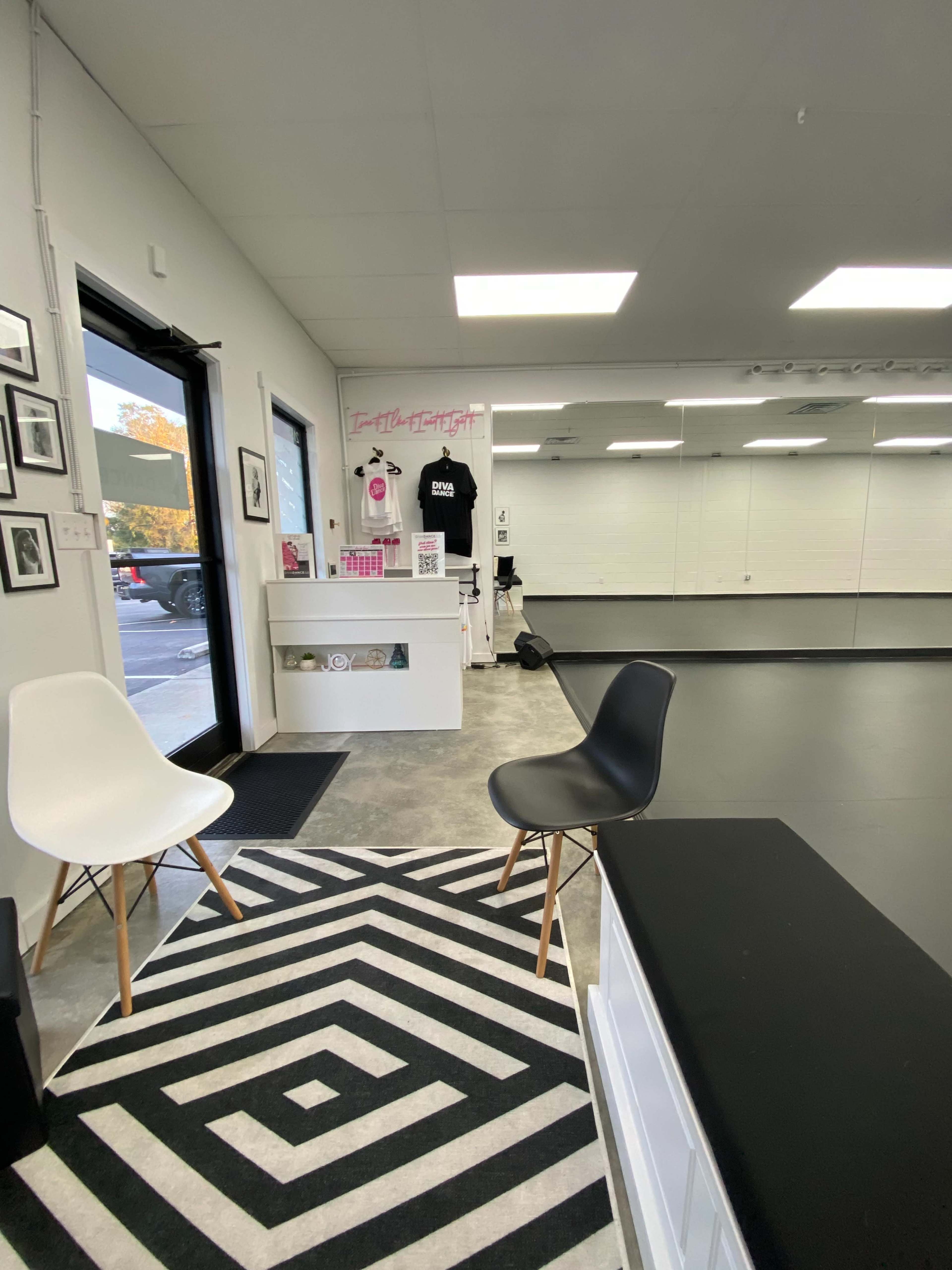 A brightly lit studio interior with two chairs on a geometric rug, a reception area displaying pink items, and a large mirrored wall.