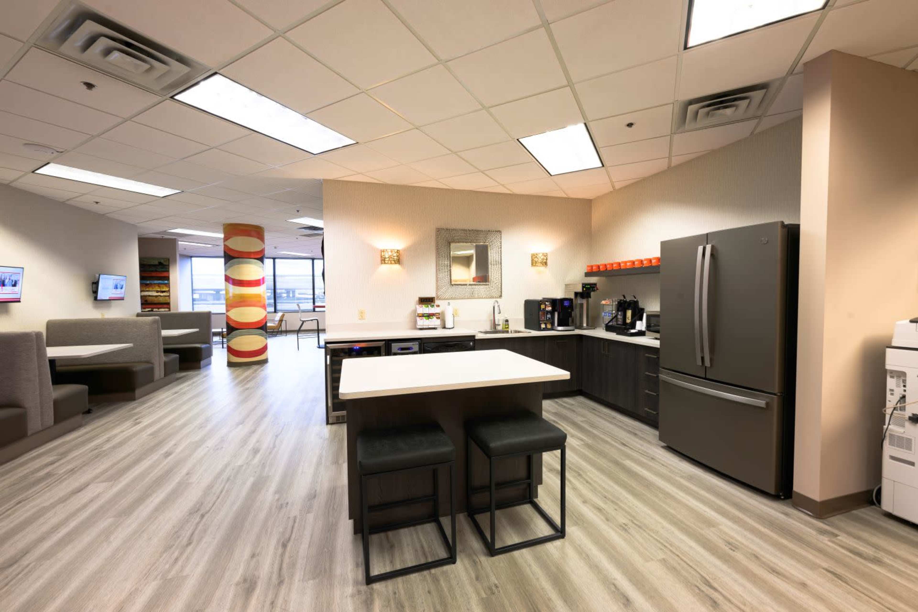 A modern office kitchen features a dark gray refrigerator, a central island with black stools, and seating areas along the walls.