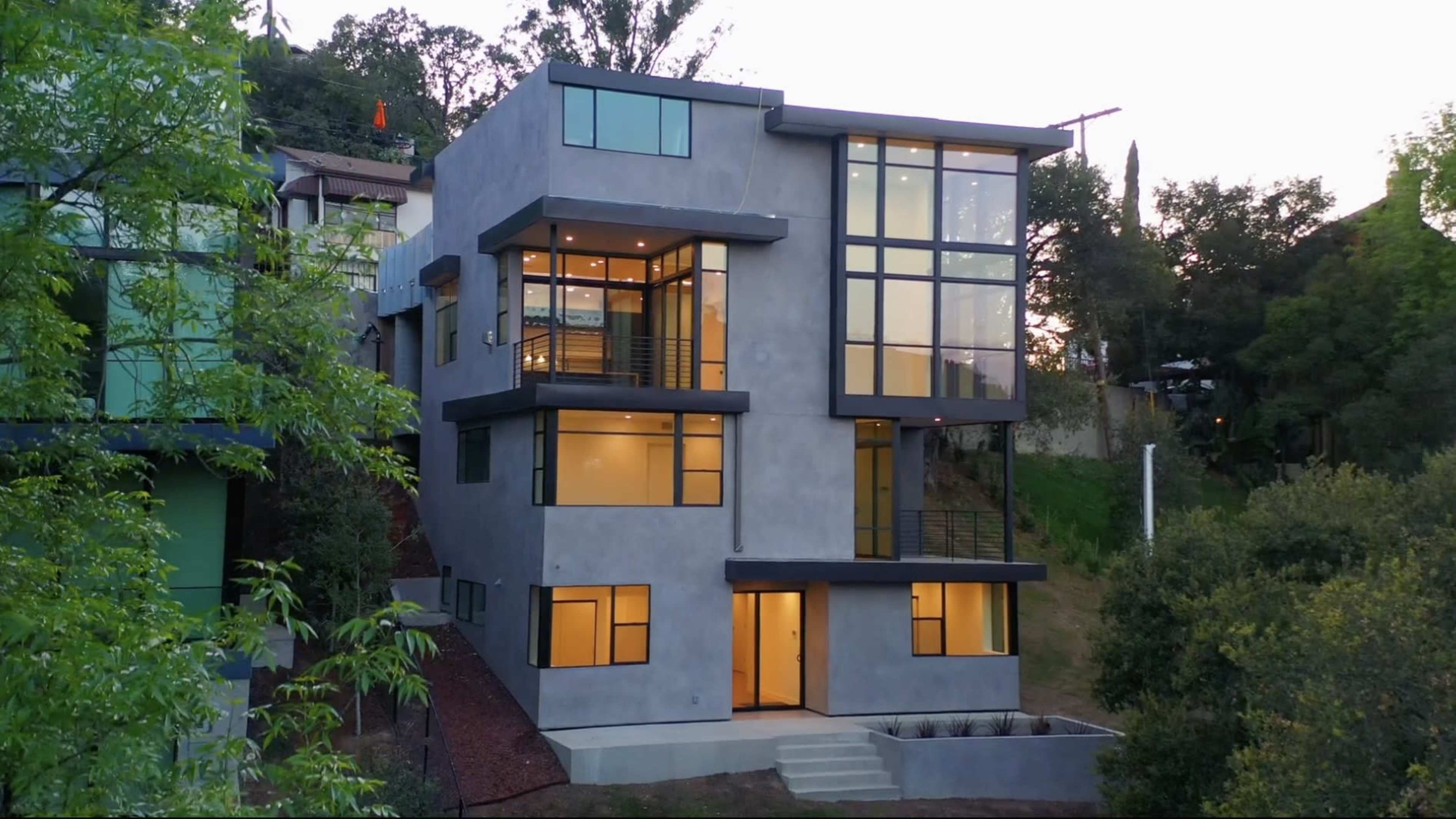 The image shows a modern multi-level house with large windows and a concrete exterior, situated on a hillside surrounded by greenery.
