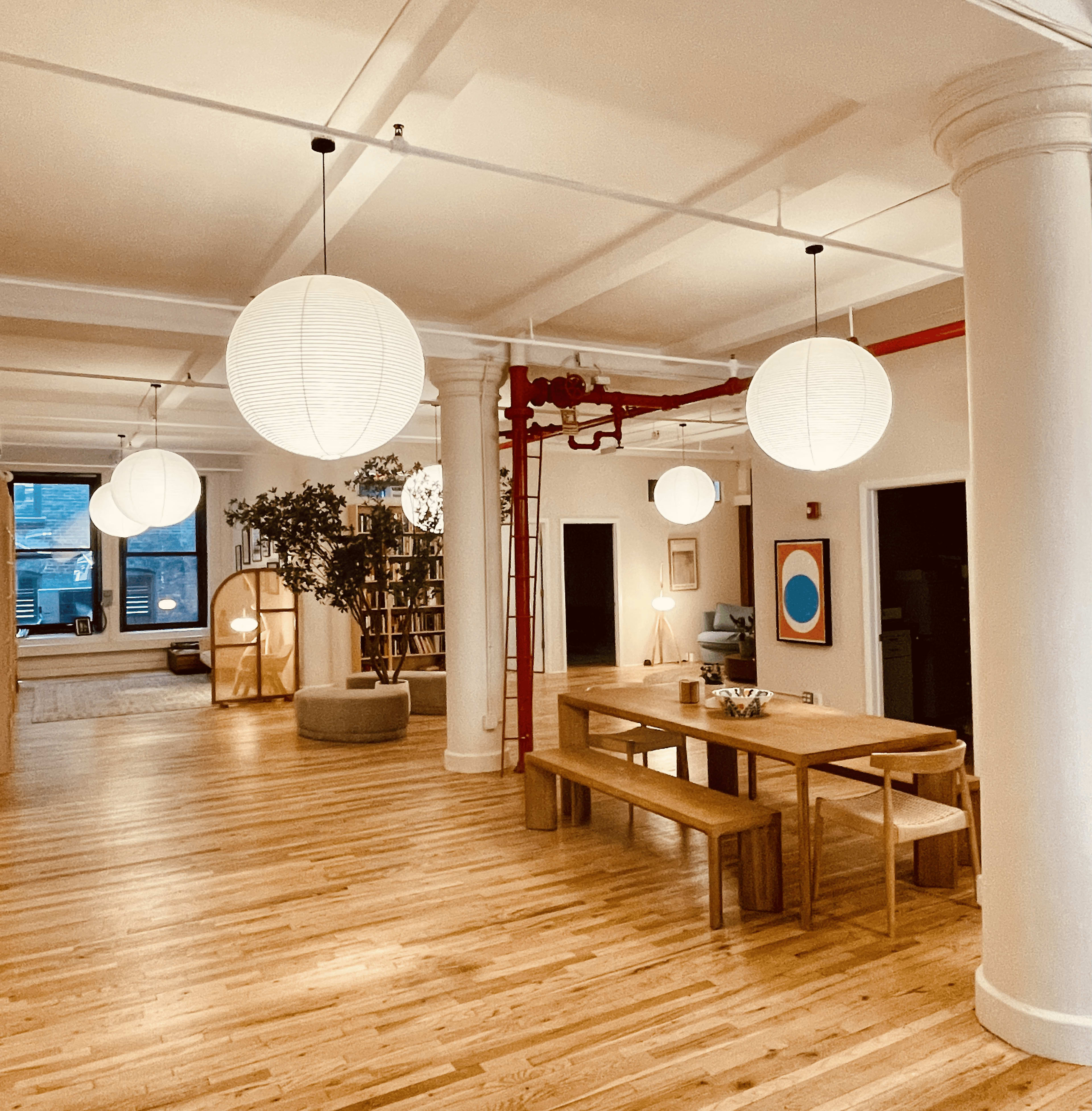 The image shows a spacious, well-lit interior with wooden floors, large pendant lamps, a long wooden table, and various decorative elements including a plant and artwork on the walls.