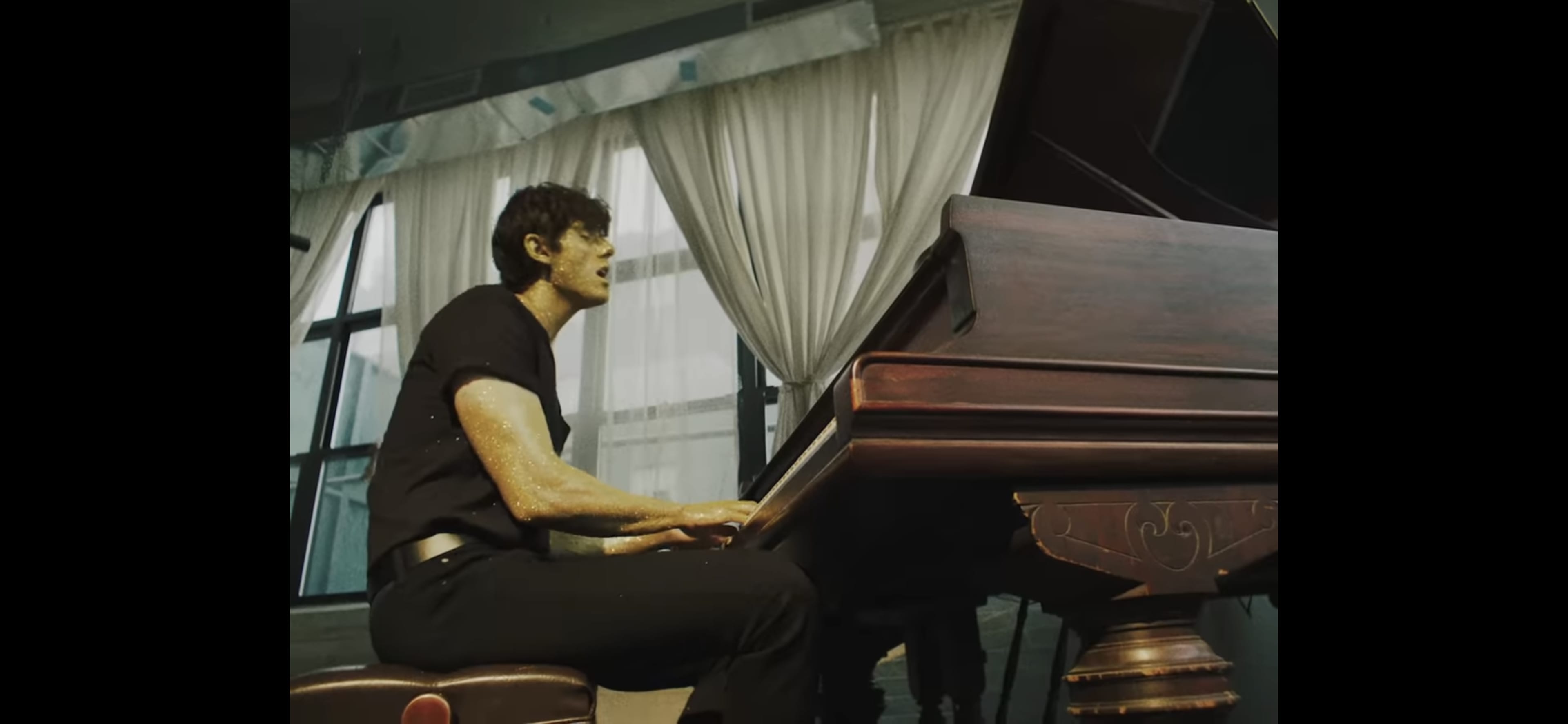 A person plays a grand piano in a room with large windows and white curtains.