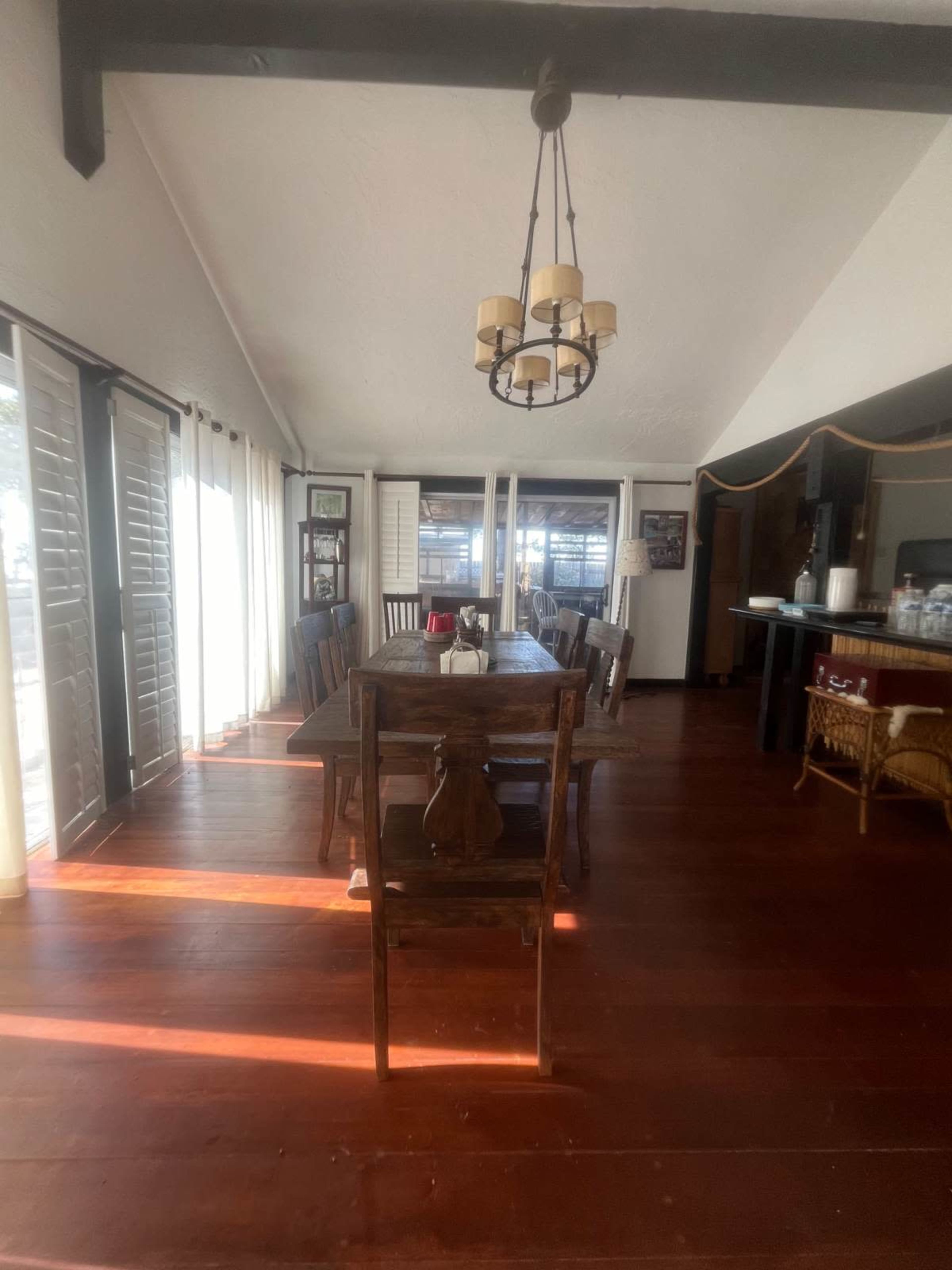 The image shows a dining area with a wooden table, chairs, and a chandelier, illuminated by natural light from large windows.