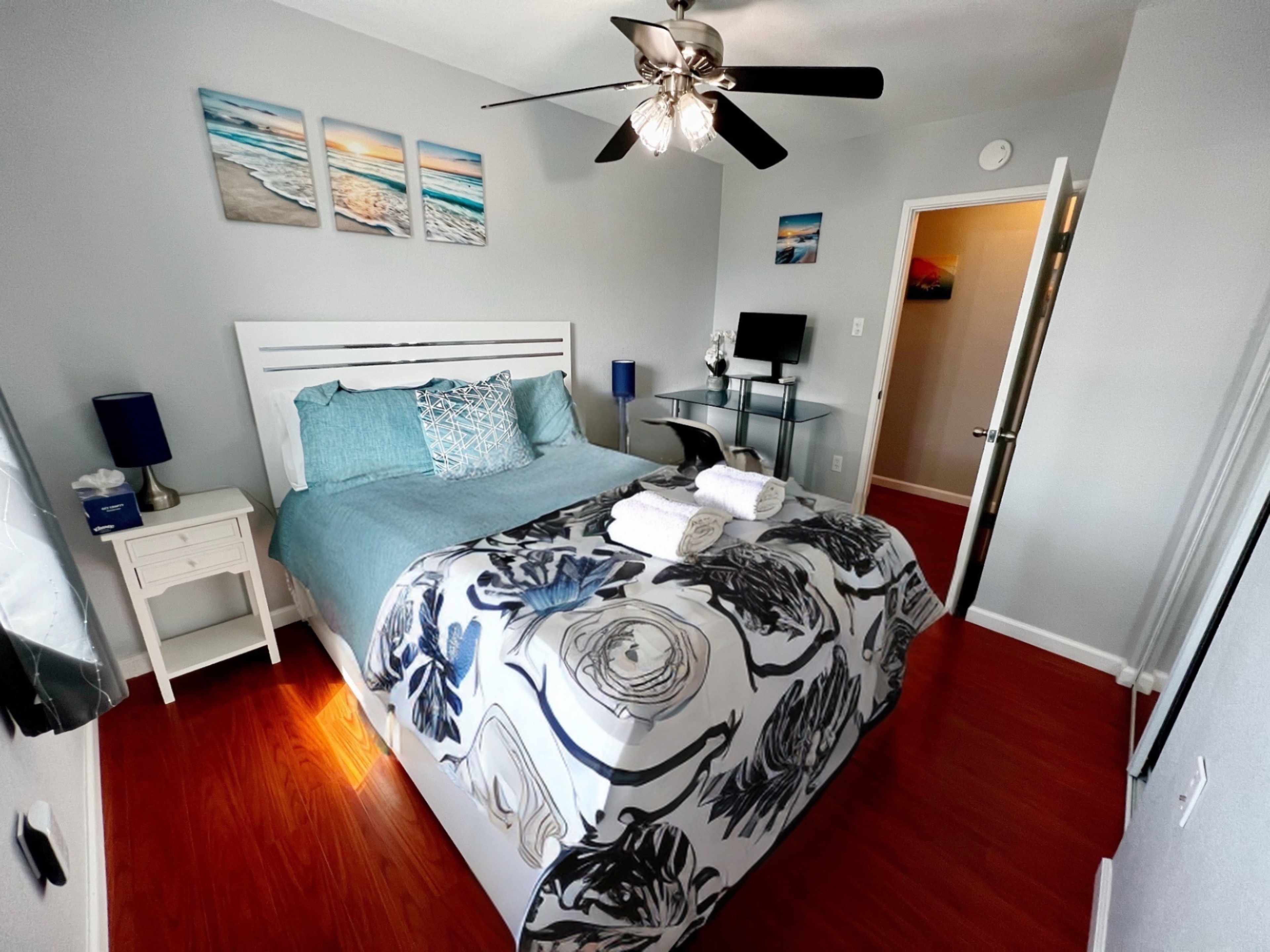 A neatly arranged bedroom featuring a bed with a floral patterned comforter, a nightstand, a desk with a chair, and a ceiling fan, all in a room with light gray walls and wooden flooring.
