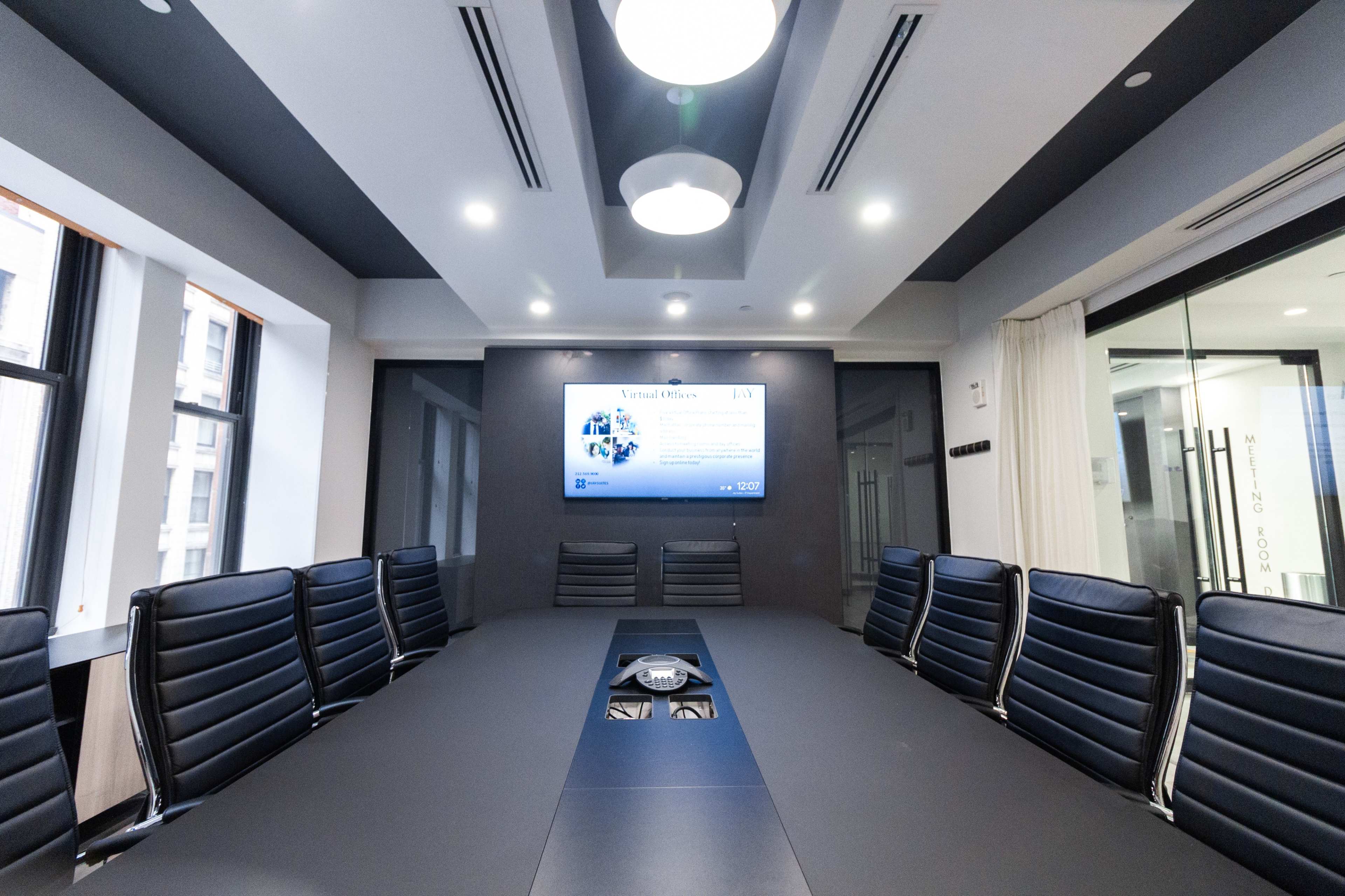 A modern conference room features a long table surrounded by black leather chairs, with a large screen displaying a presentation on the wall.