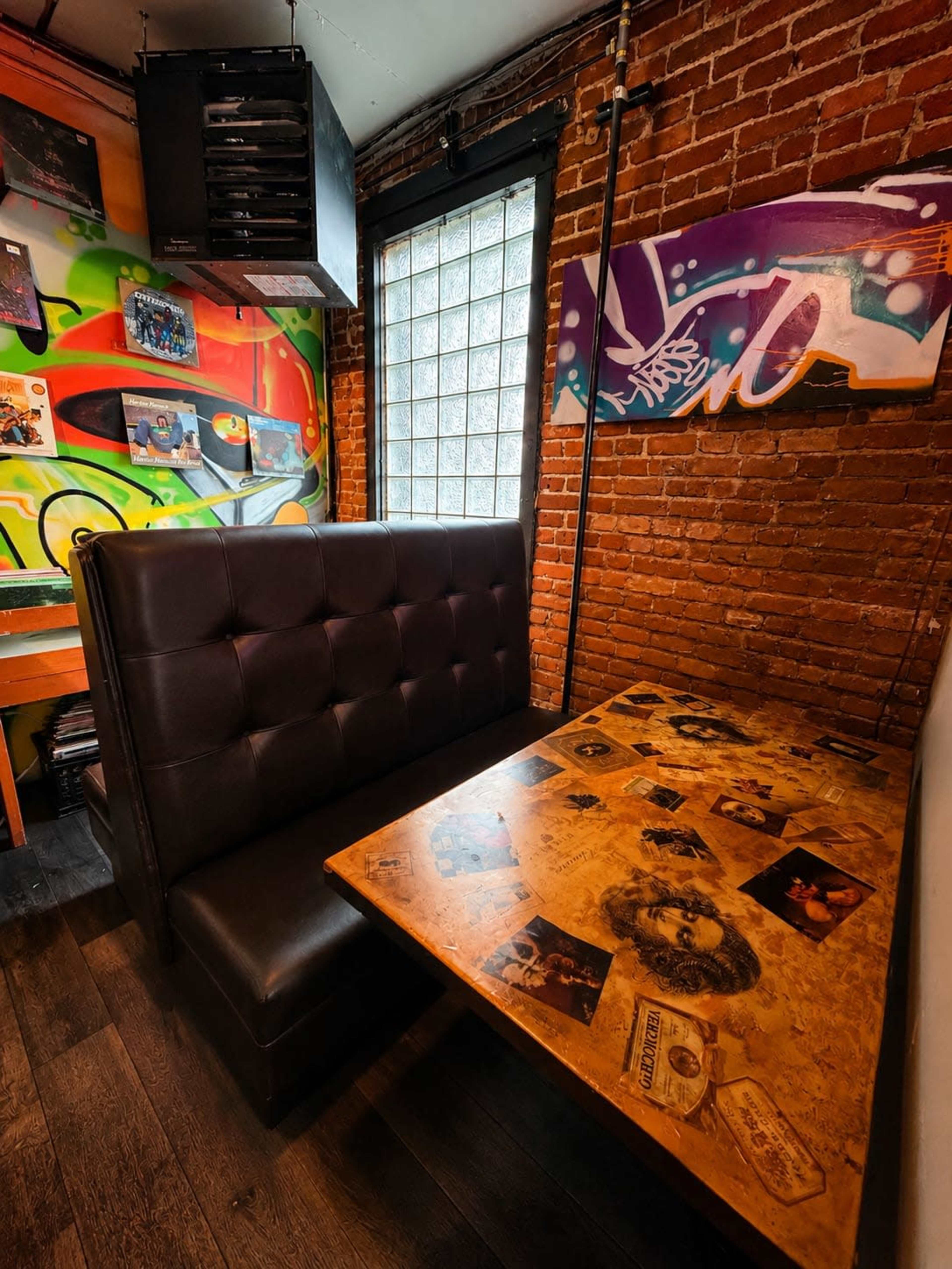 The image shows a cozy booth in a restaurant with a wooden table decorated with various photographs and artwork, against a backdrop of colorful graffiti on a brick wall.