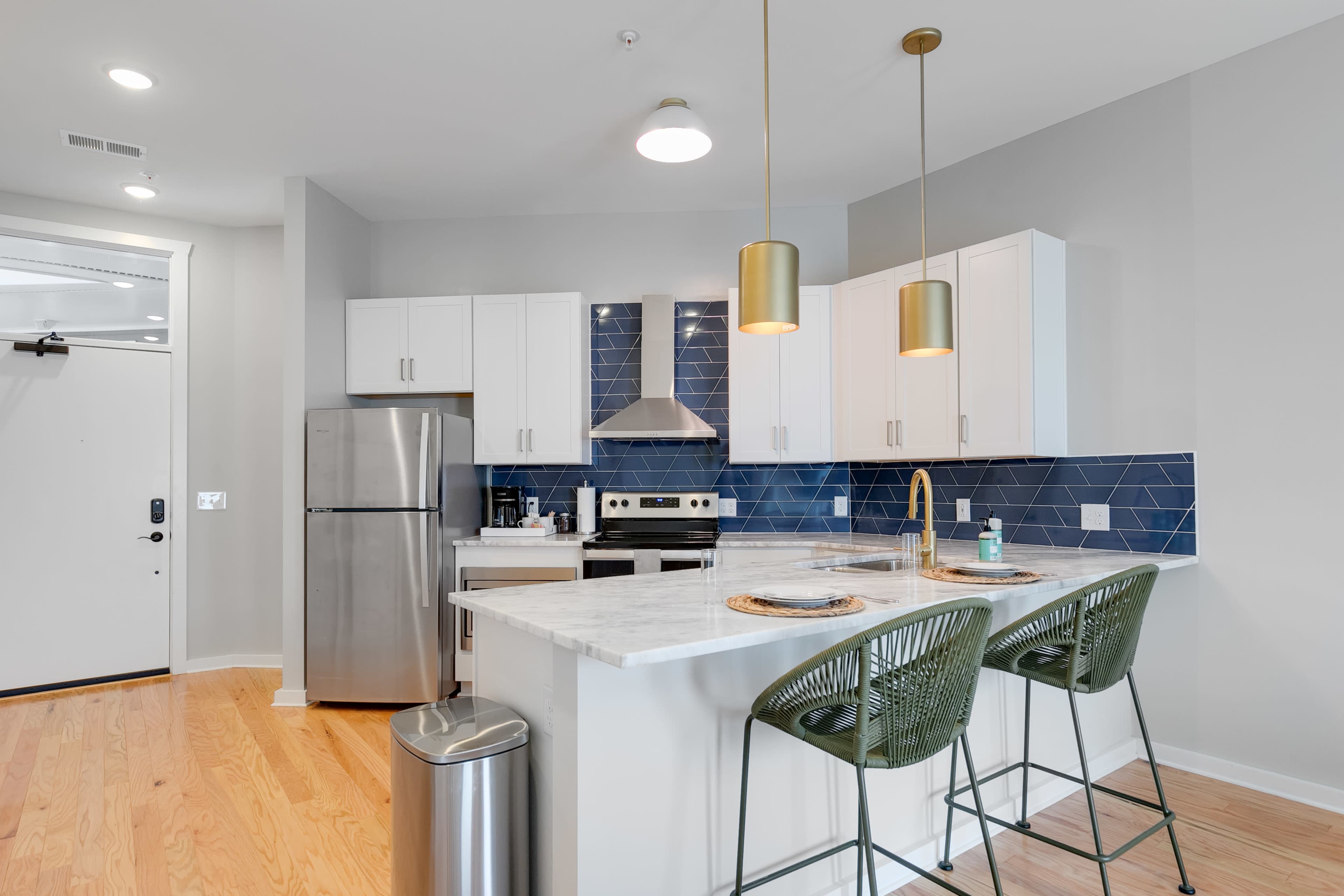 A modern kitchen with stainless steel appliances, white cabinets, a marble countertop, and green bar stools.