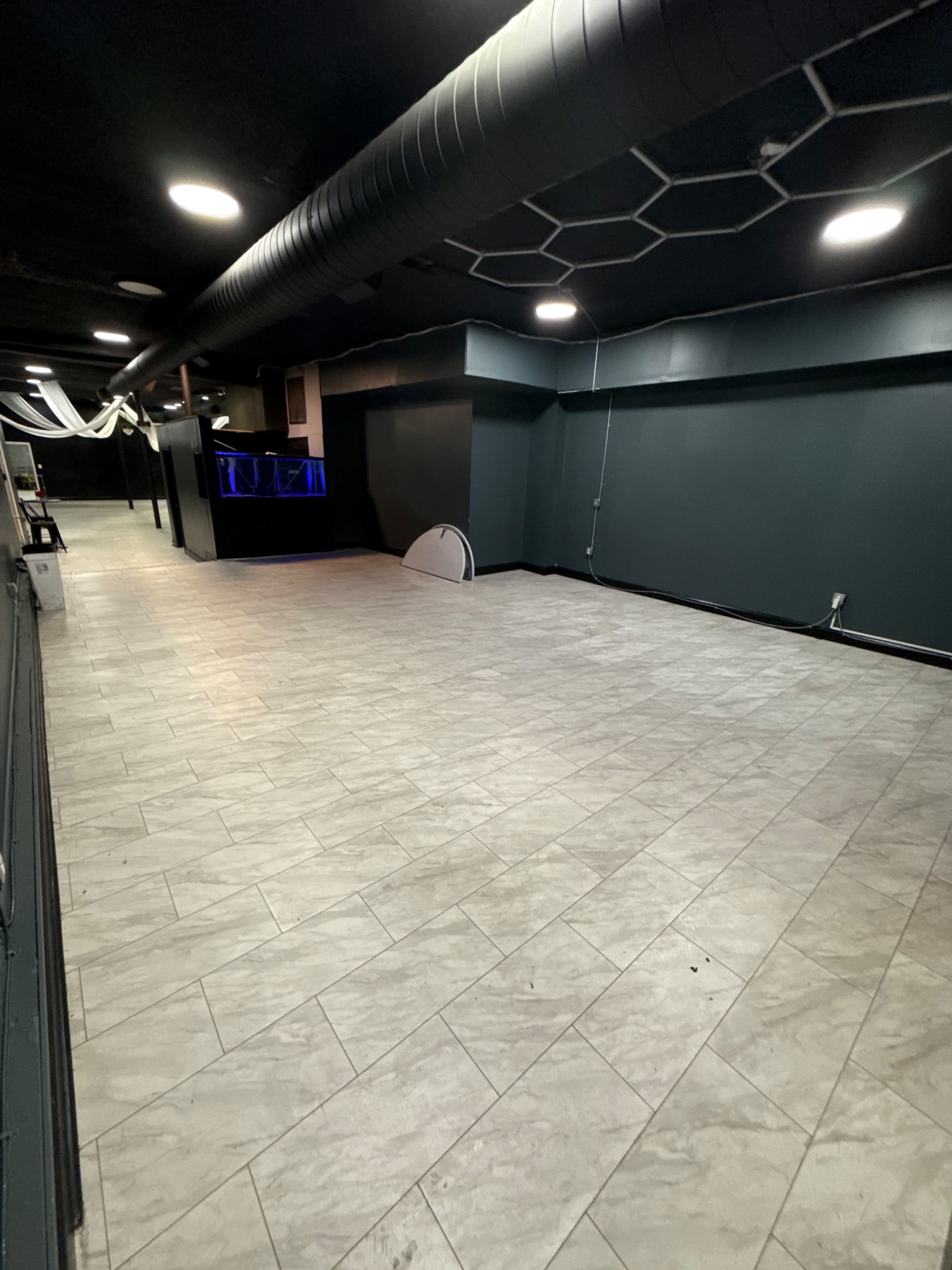 The image shows an empty interior space with grey tiled flooring, dark walls, and a bar area illuminated by blue lighting in the background.
