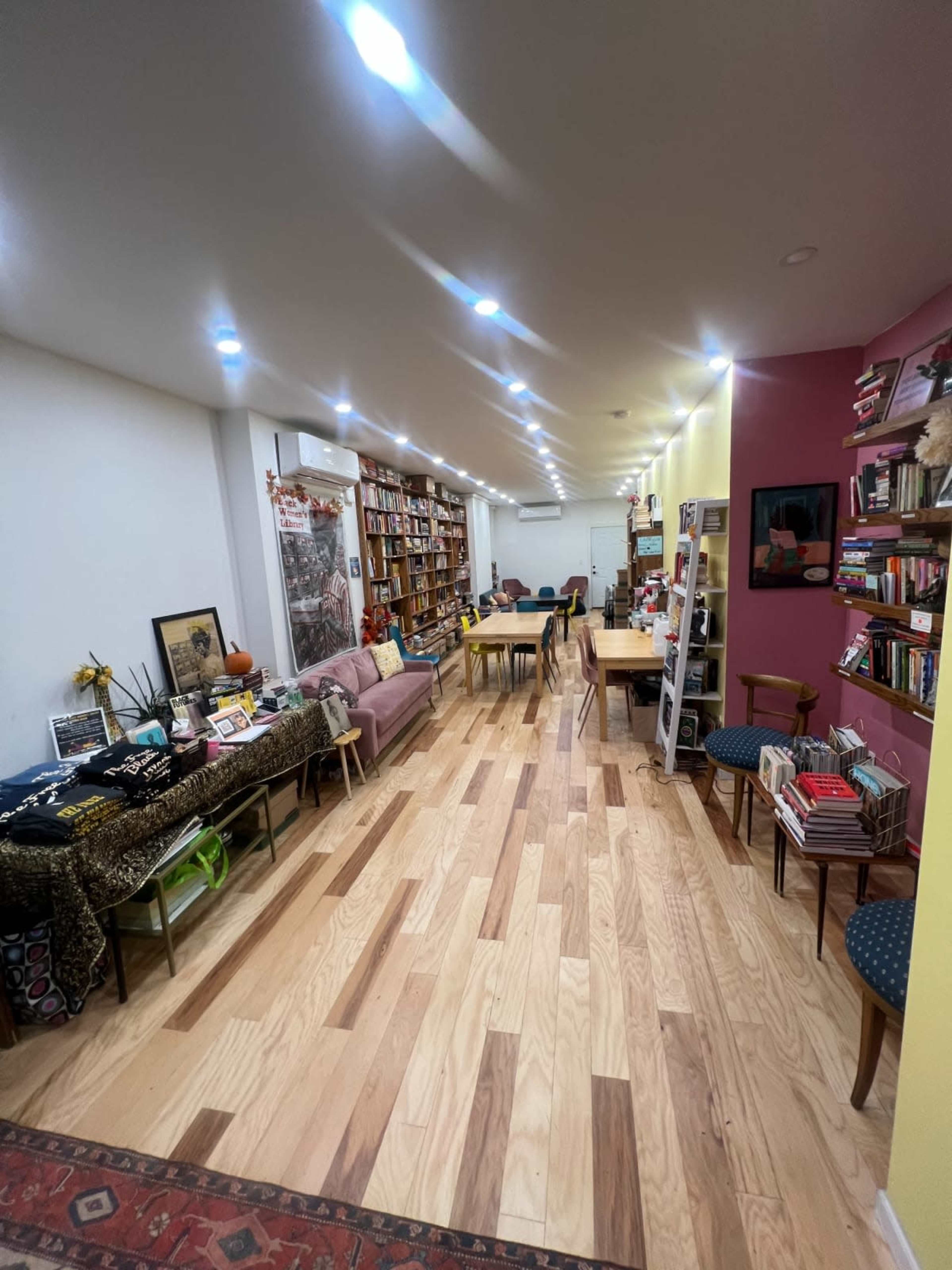 The image shows a long, narrow room lined with bookshelves on one side and a seating area on the other, featuring wooden floors and various reading materials.