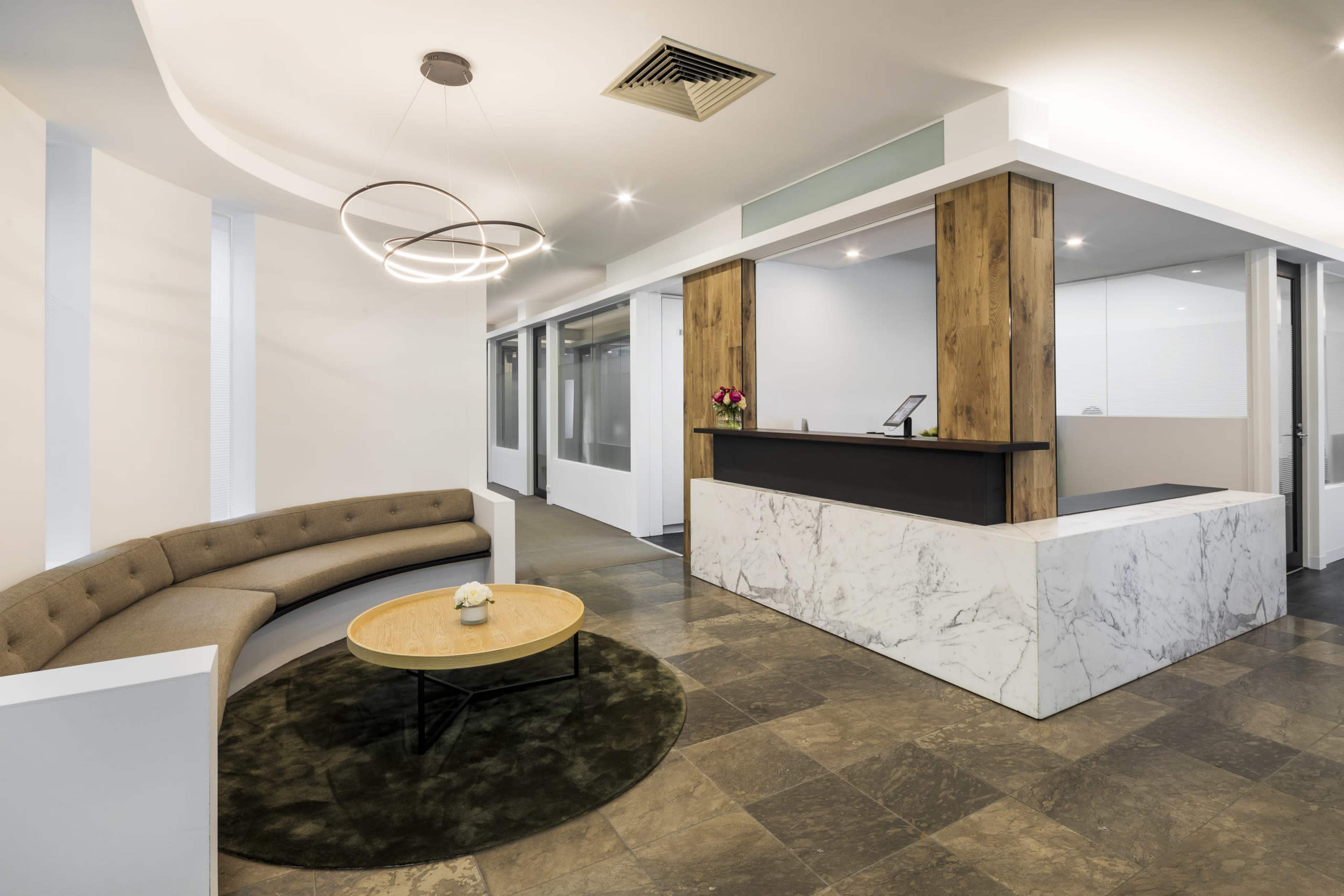 The image shows a modern reception area featuring a curved sofa, a round coffee table, and a front desk made of wood and marble.
