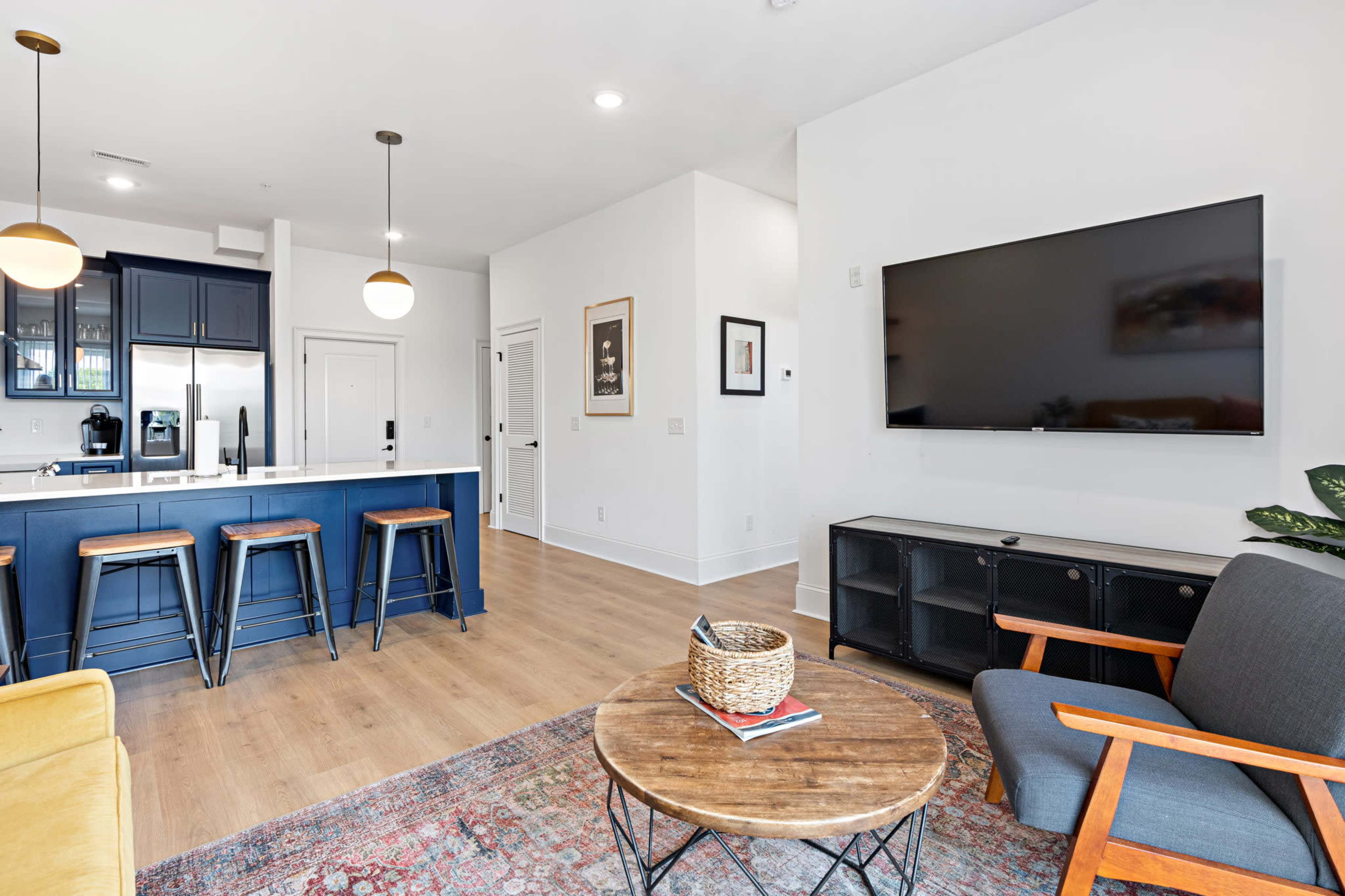 The image shows a modern living space featuring a kitchen bar with three stools, a large television mounted on the wall, and a seating area with a round coffee table and a plant.