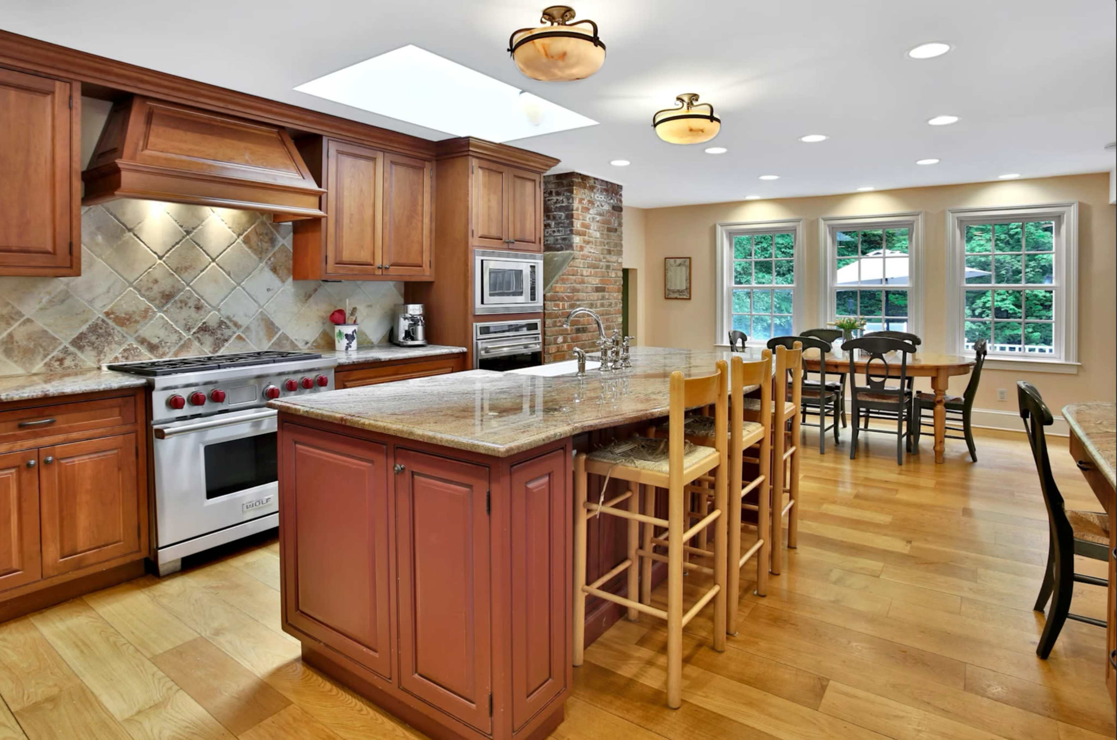 The image shows a modern kitchen with wooden cabinetry, a large island with bar stools, and a dining area featuring a table and chairs, all well-lit by natural light.