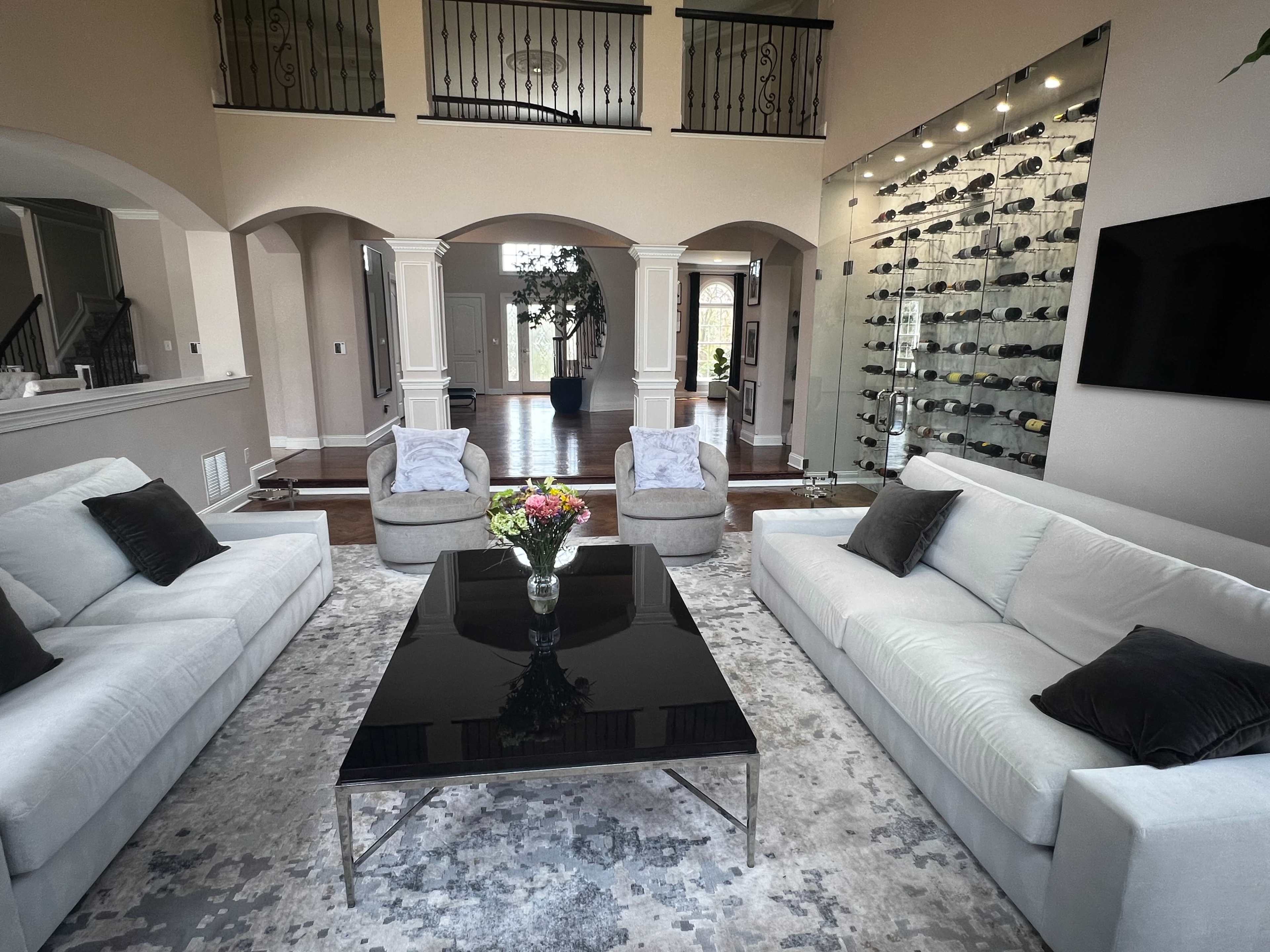 The scene features a spacious living area with a black coffee table at its center, two light gray sofas, two armchairs, and a wall displaying an assortment of wine bottles.
