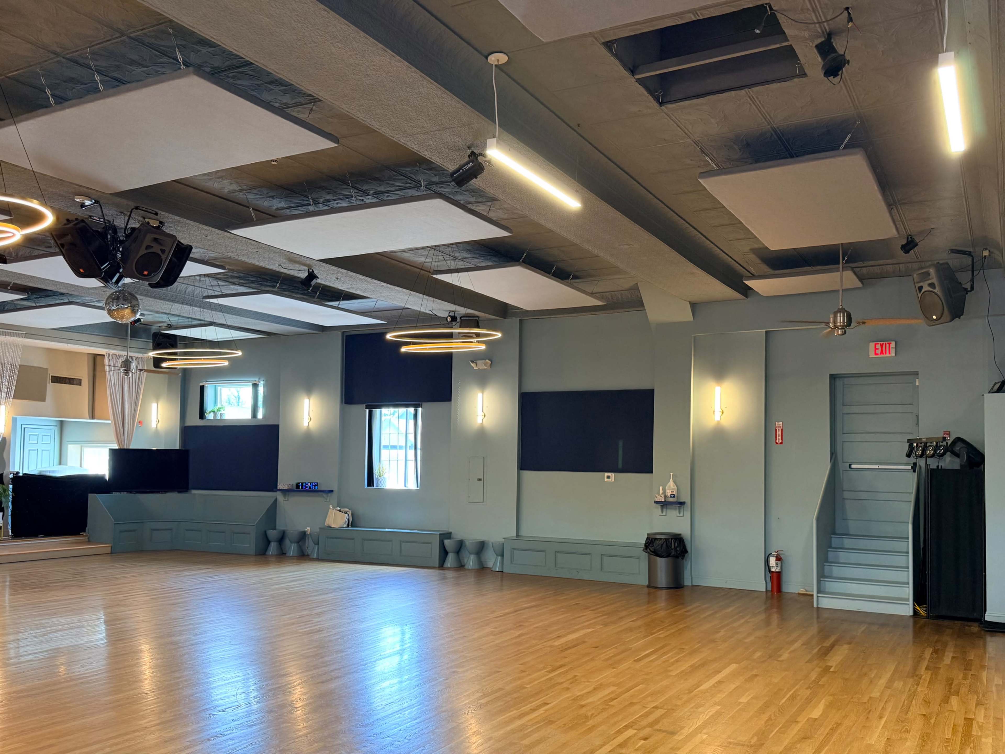 The image shows an empty dance studio with a polished wooden floor, gray walls, and modern lighting fixtures.