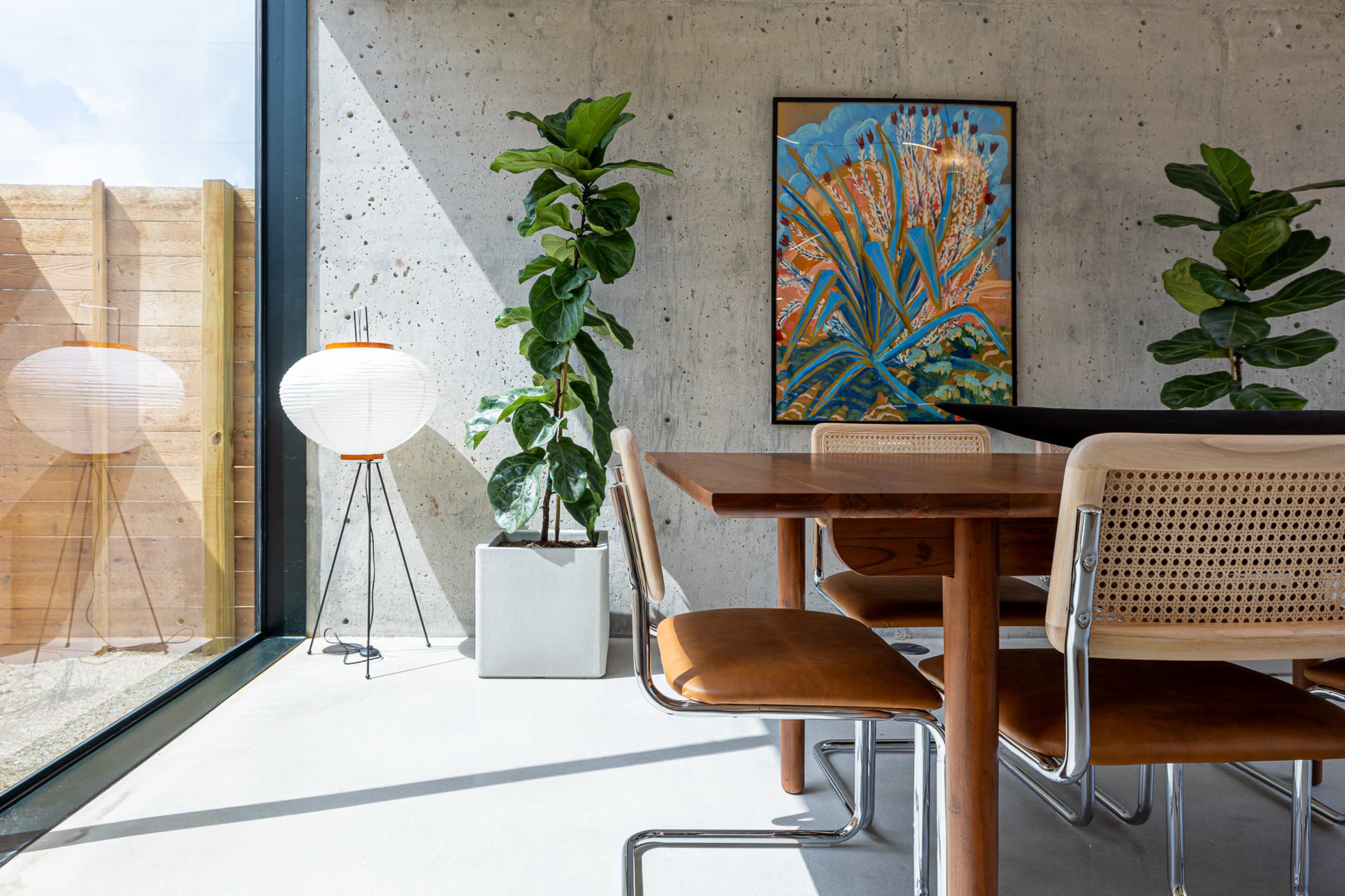 A wooden dining table is set in a modern interior with concrete walls, surrounded by chairs and adorned with plants, while a colorful artwork hangs on the wall.