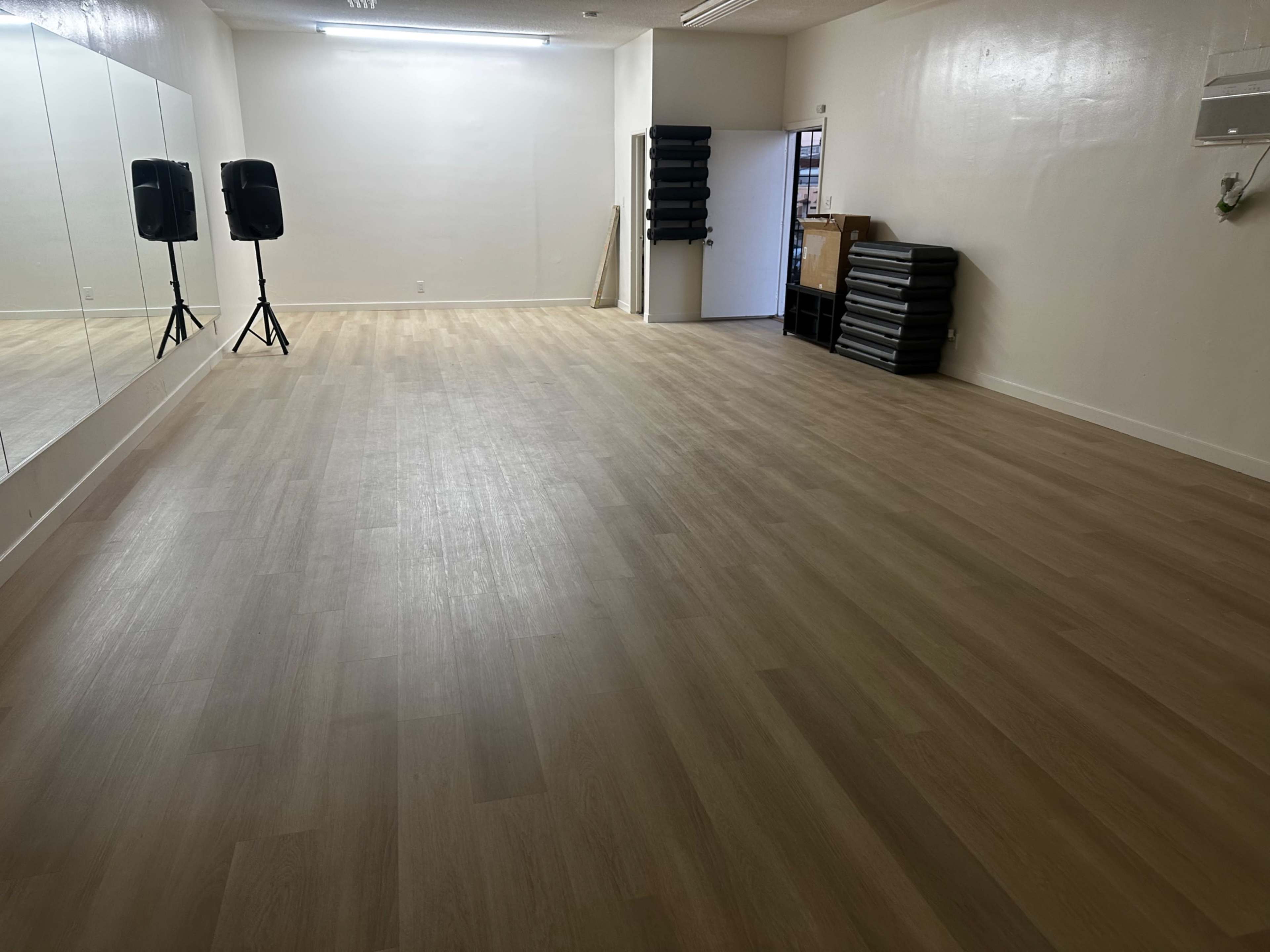 The image shows a spacious, empty studio with light wooden flooring, mirrored walls, and sound equipment on stands.