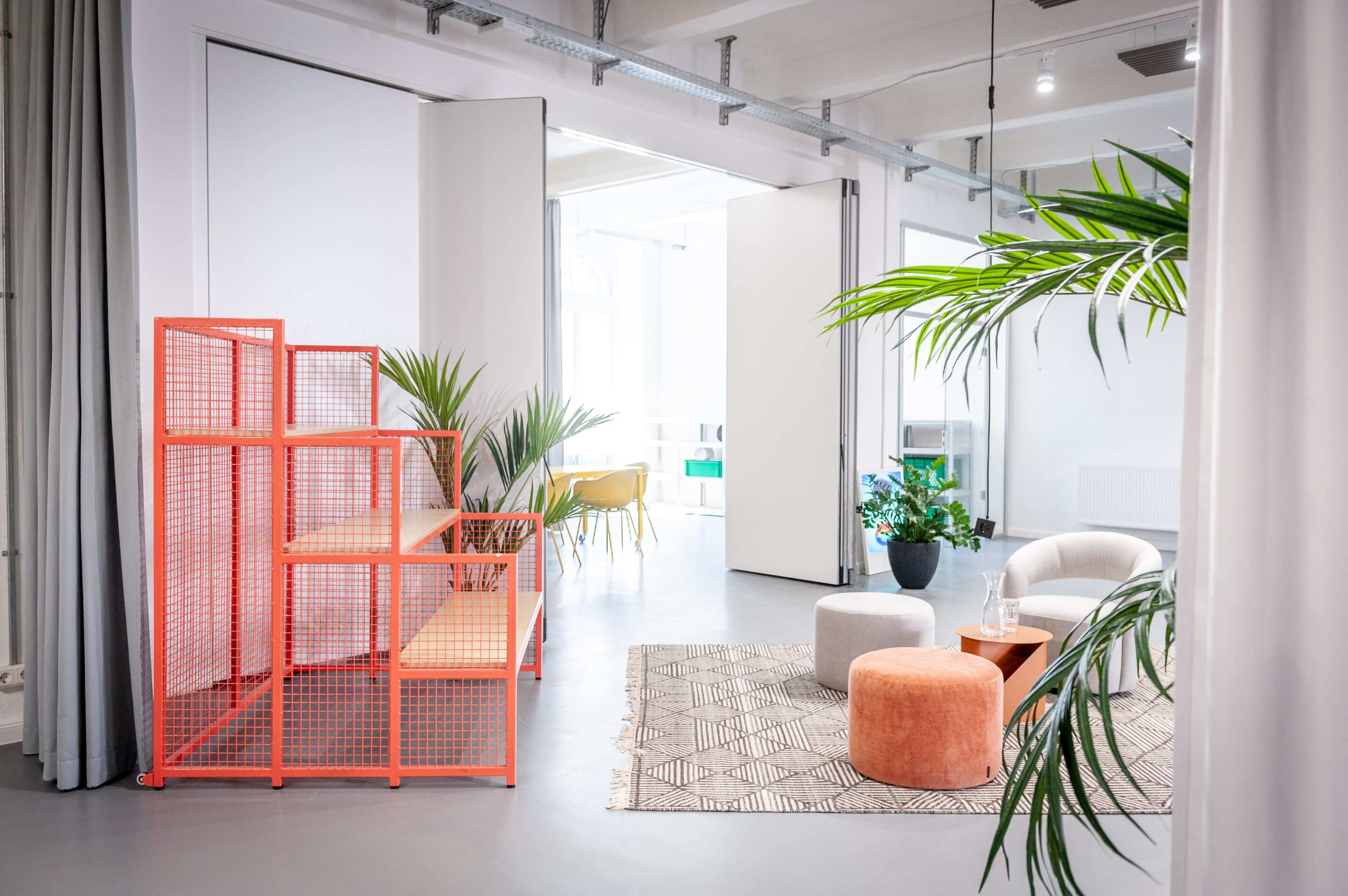 A modern interior space with an orange wire shelf, a plant, and various seating options, including round poufs, arranged on a patterned rug.