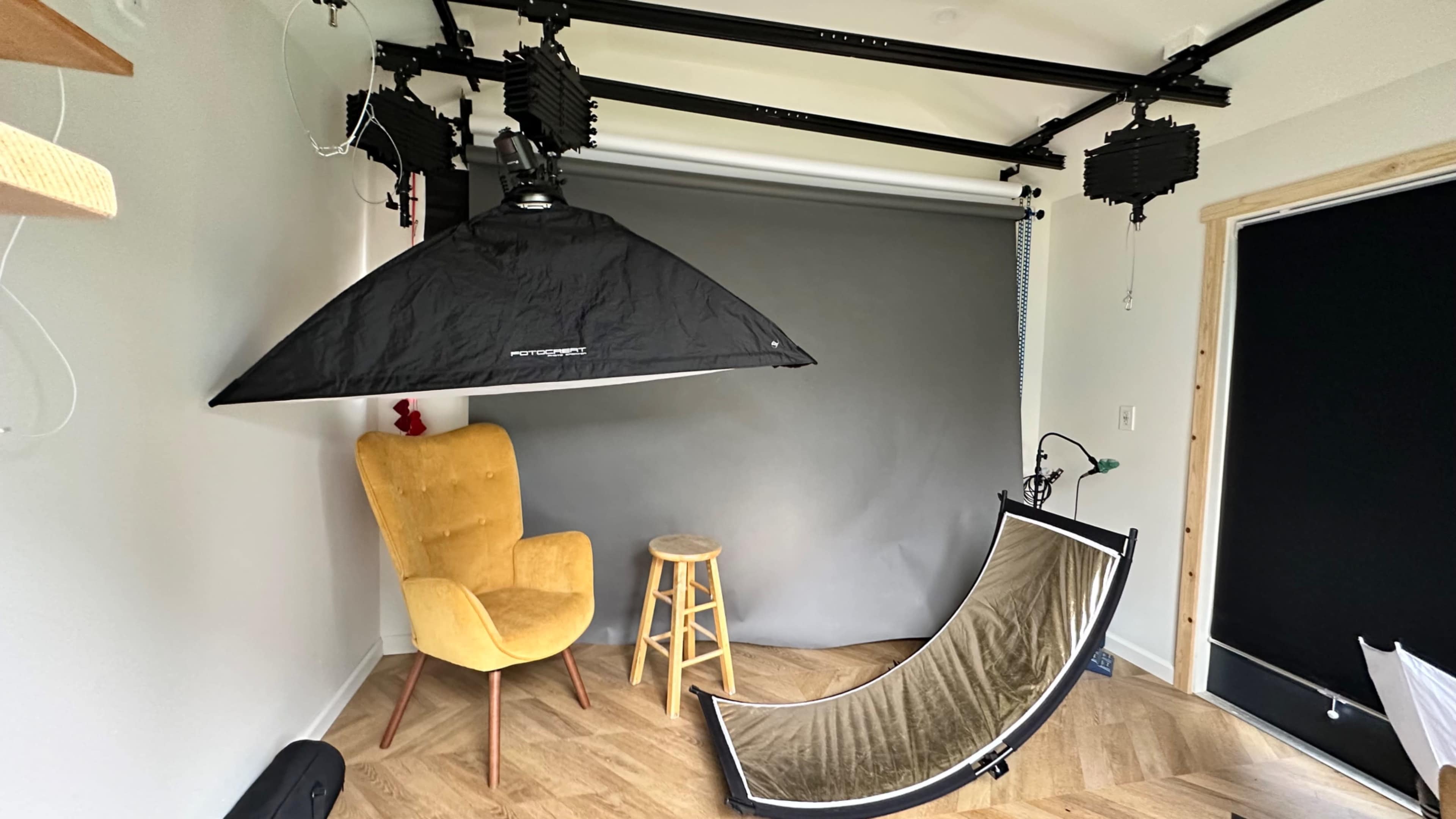A photography studio setup featuring a gray backdrop, a yellow chair, a wooden stool, and various lighting equipment.