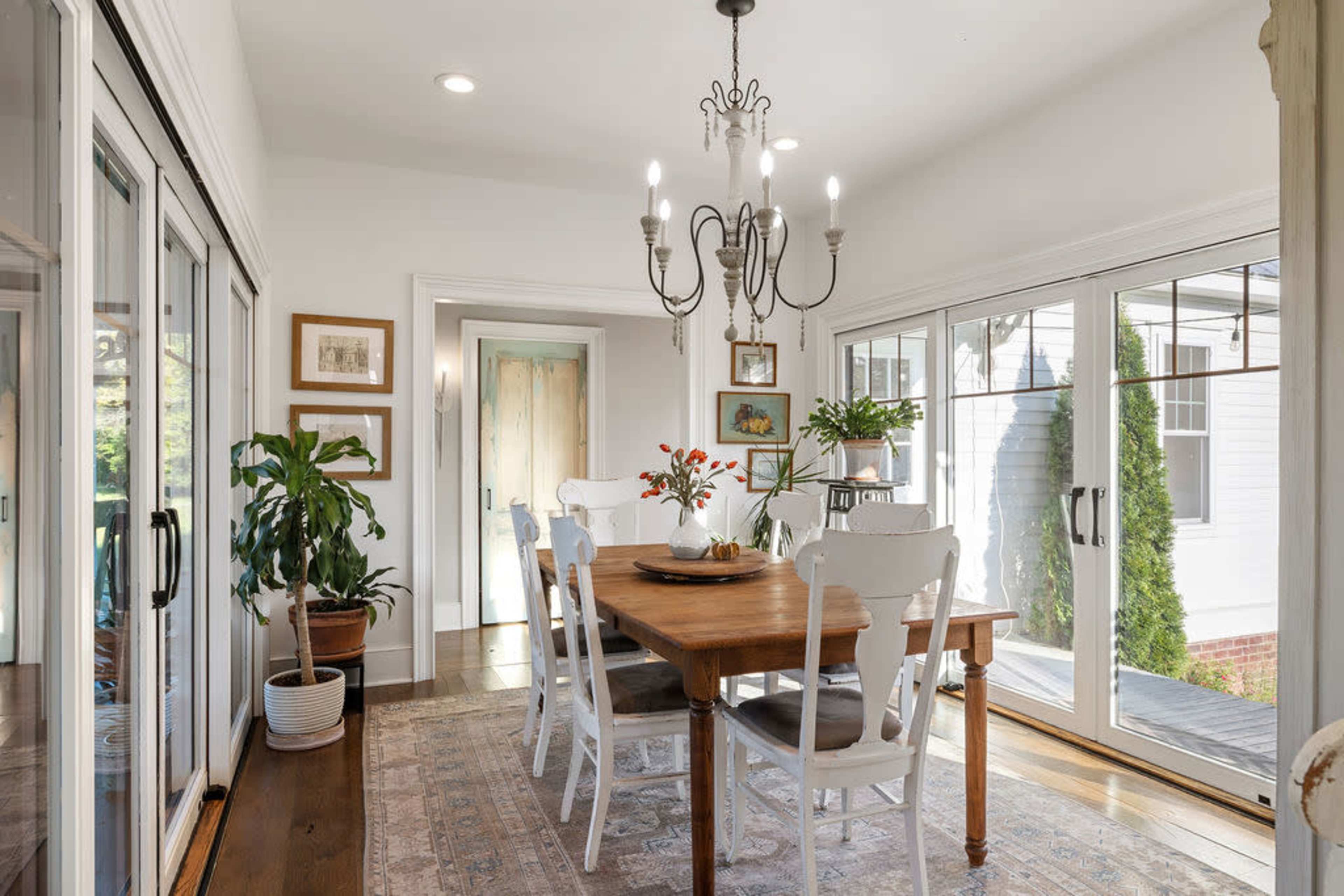 A well-lit dining room features a wooden table surrounded by white chairs, with large glass doors providing a view of the outdoors.