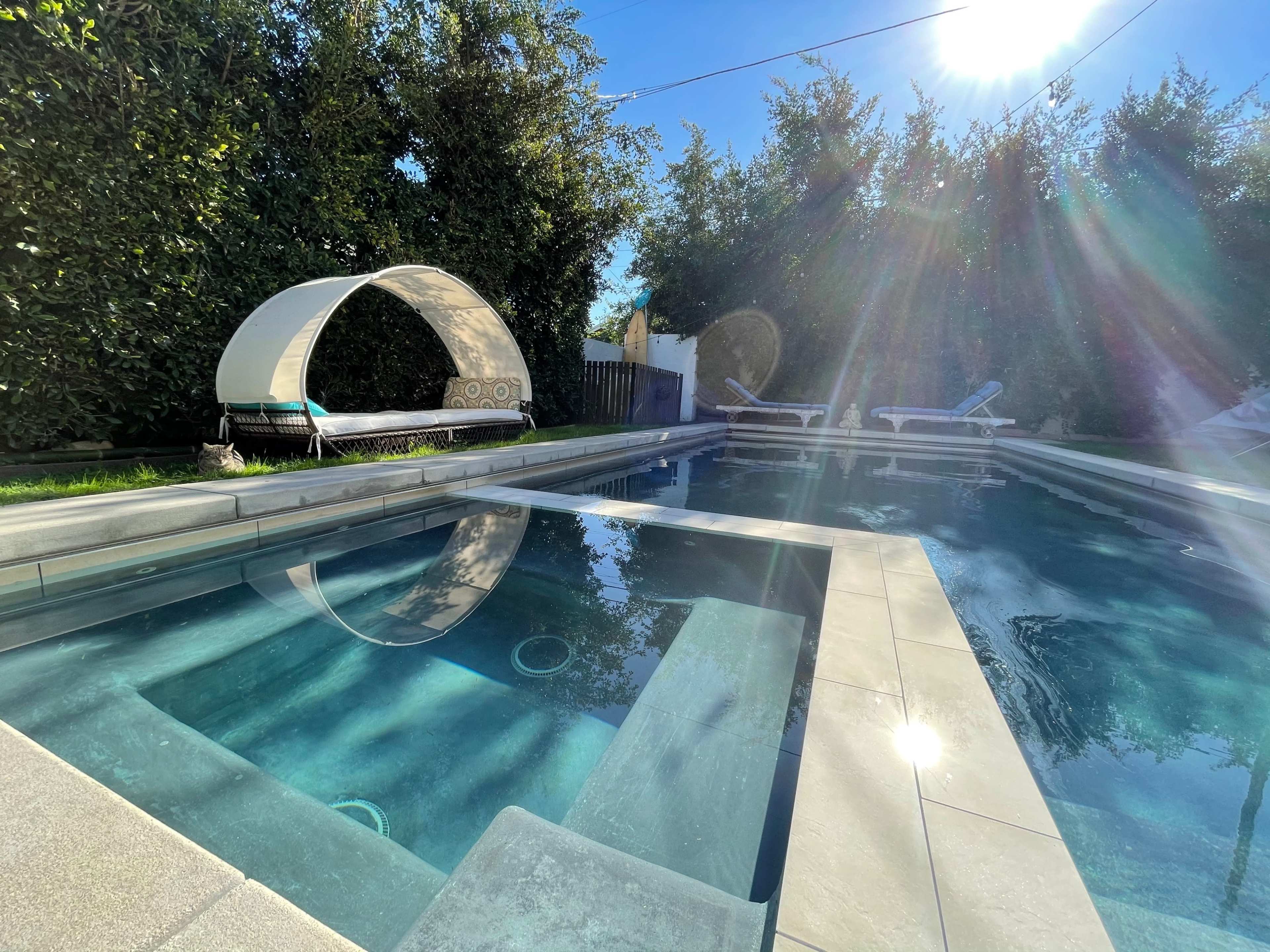 A swimming pool surrounded by greenery, featuring a lounge area with a canopy and several deck chairs near the water.