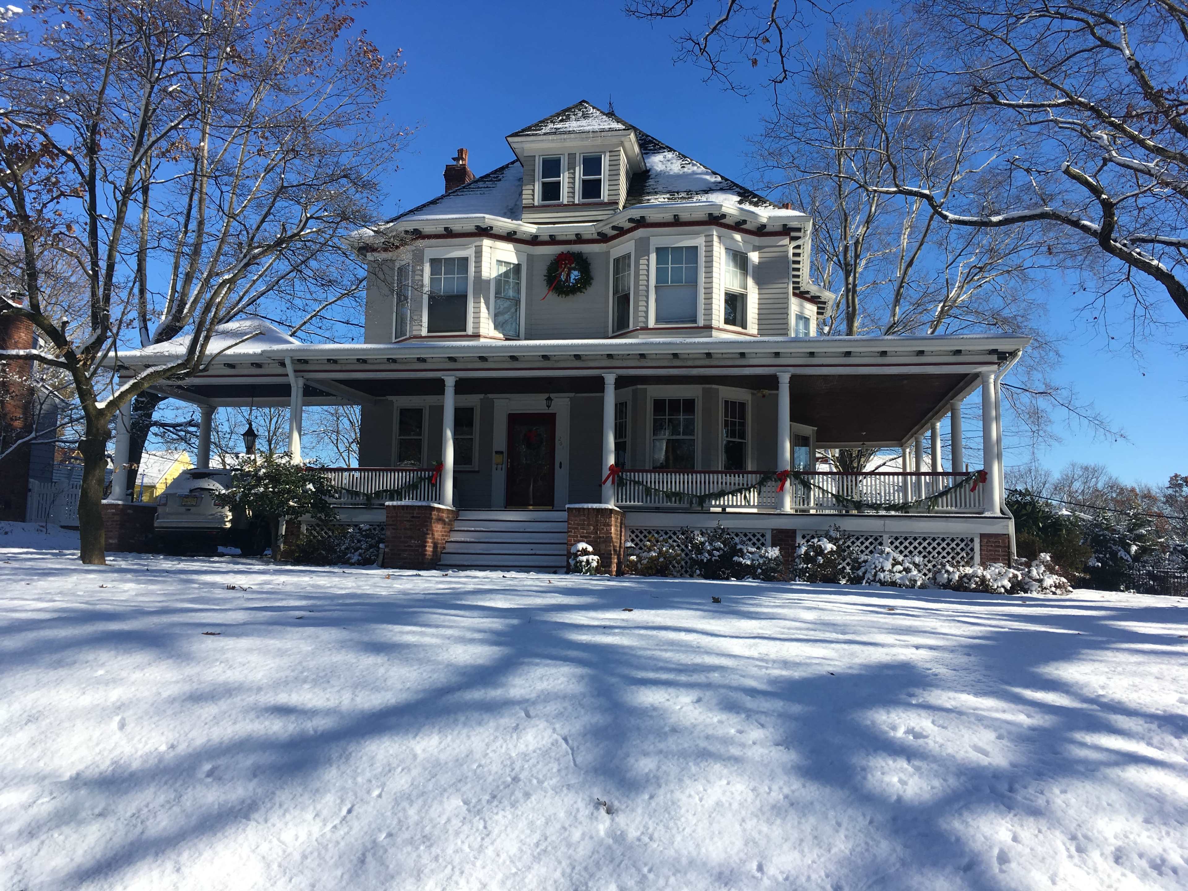 A large, two-story house with a porch is surrounded by snow-covered ground and trees on a clear winter day.