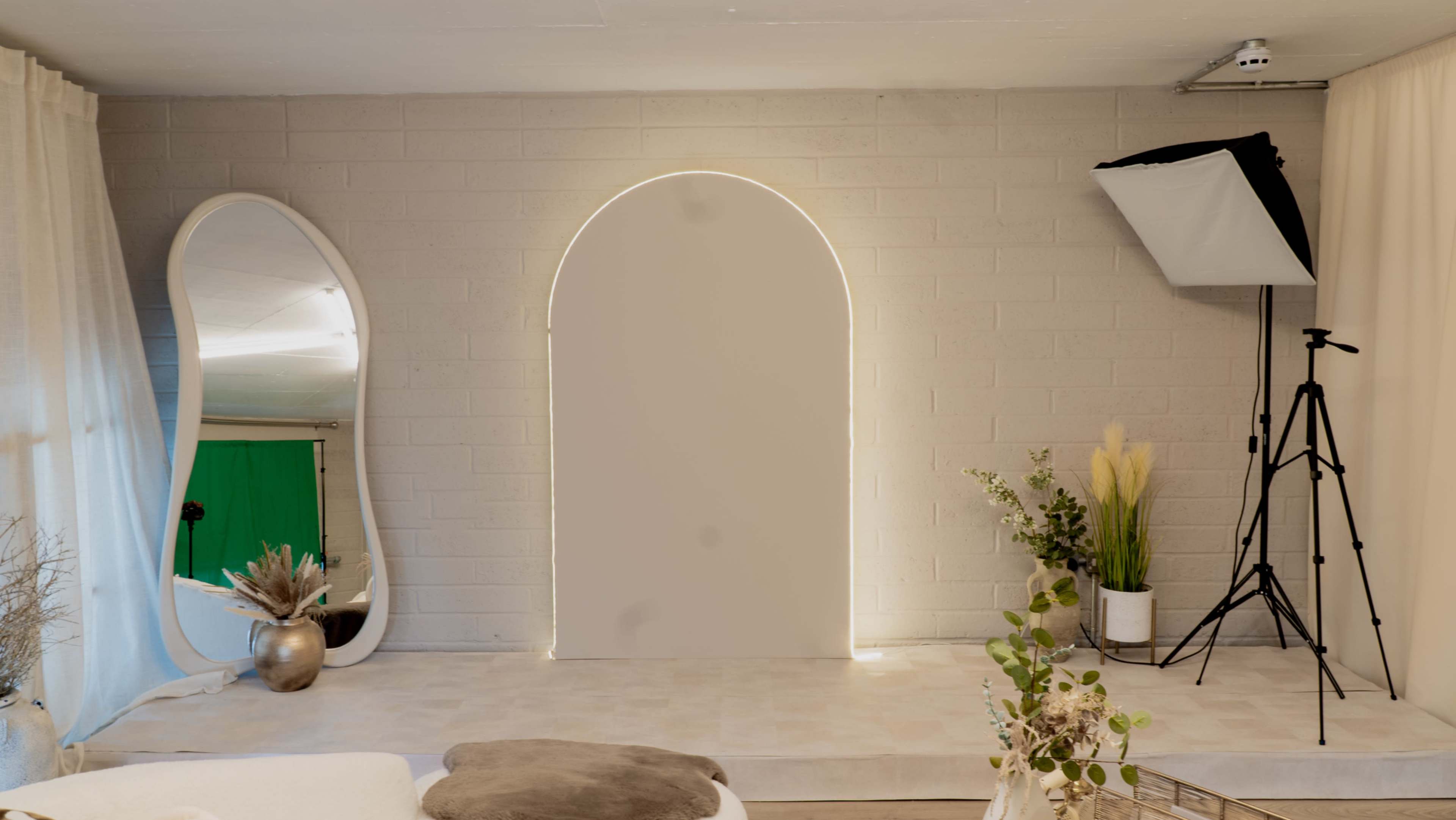The image shows a minimalist studio space with an arched backdrop illuminated by light, a large mirror, a tripod with a softbox, and decorative plants.