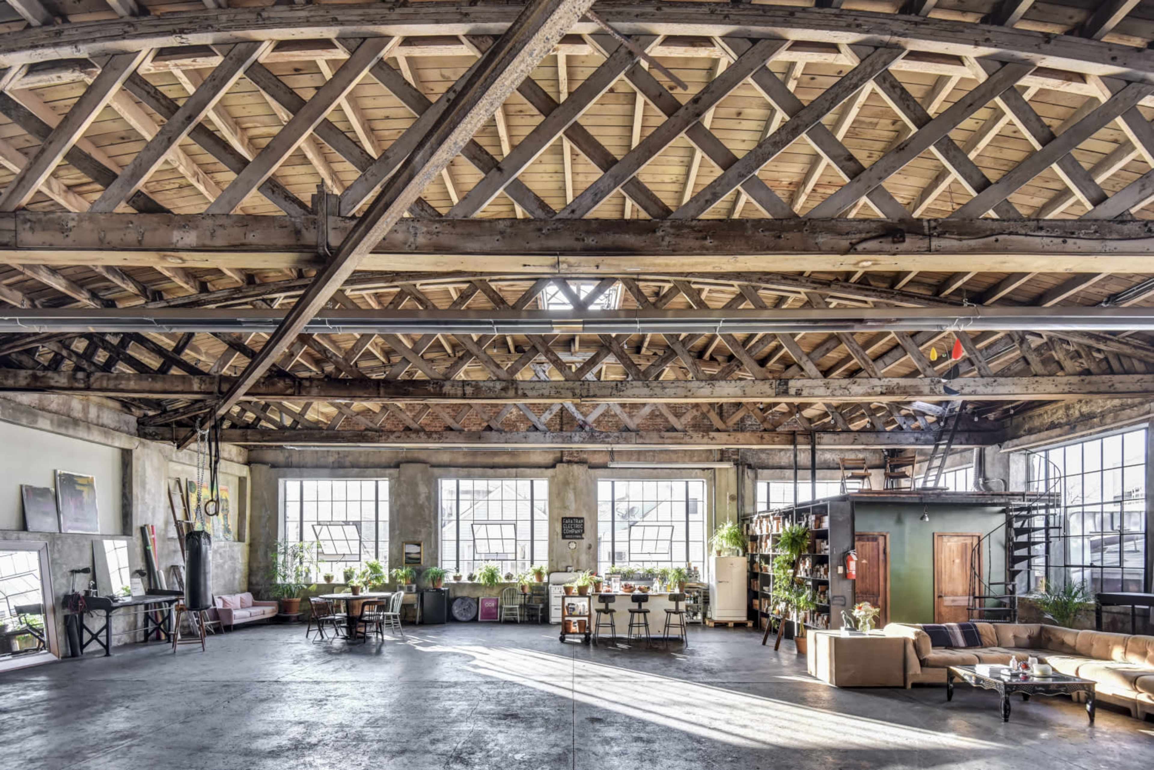 A spacious industrial-style loft with high wooden beams, large windows, and various seating areas filled with plants.